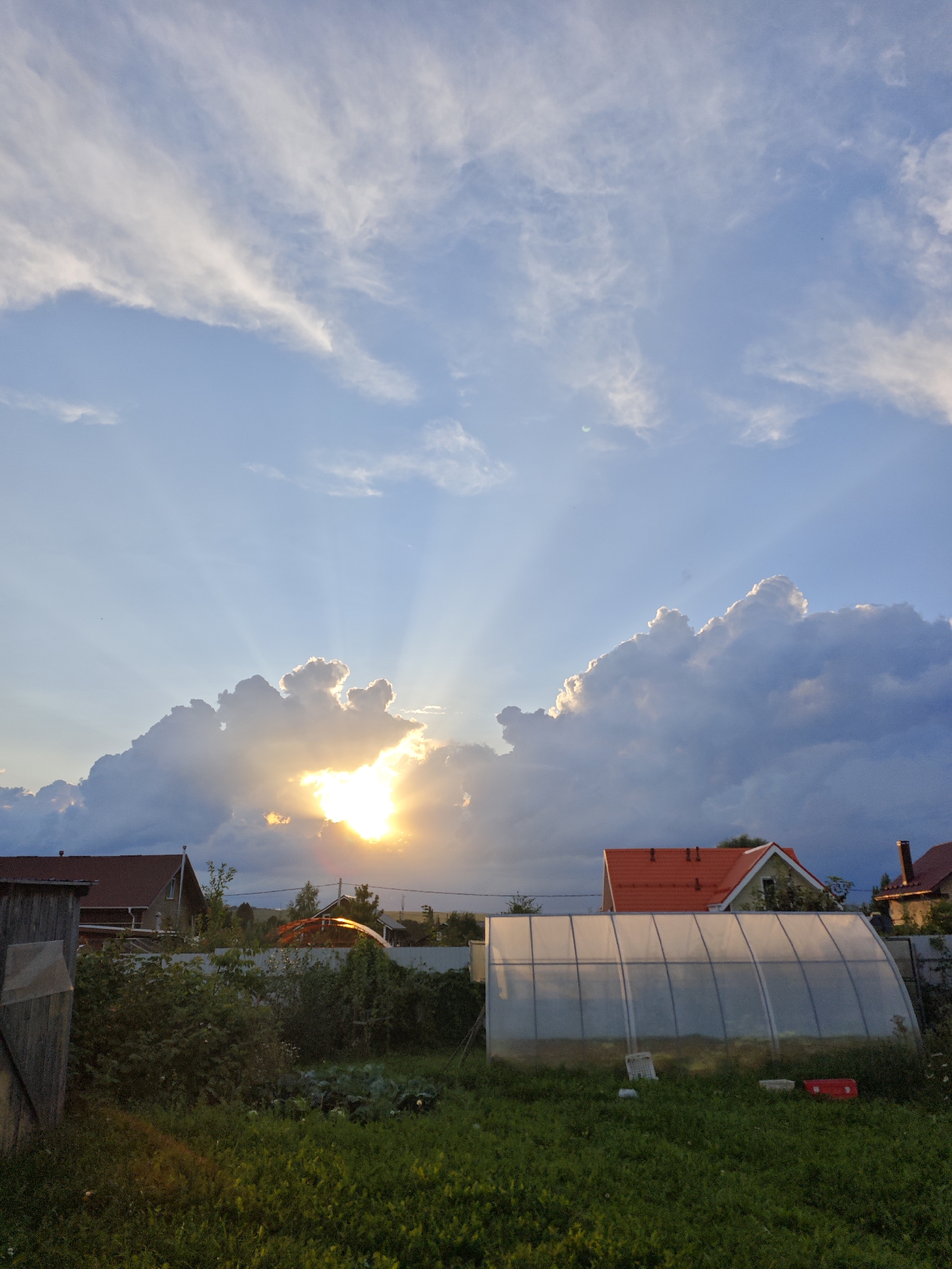The image depicts a rural scene at sunset in the city of Mokino, Russia. The sky is dominated by a dramatic display of clouds, with the sun peeking through a large cumulus formation, casting visible crepuscular rays. In the foreground, there is a green lawn with various types of vegetation. A translucent greenhouse structure is visible, with a red roof of a nearby house partially obscured by it. Further back, another house with a red roof and a chimney is visible. A wooden shed or fence is present on the far left of the image. The overall impression is one of a serene, pastoral evening in a dacha settlement.