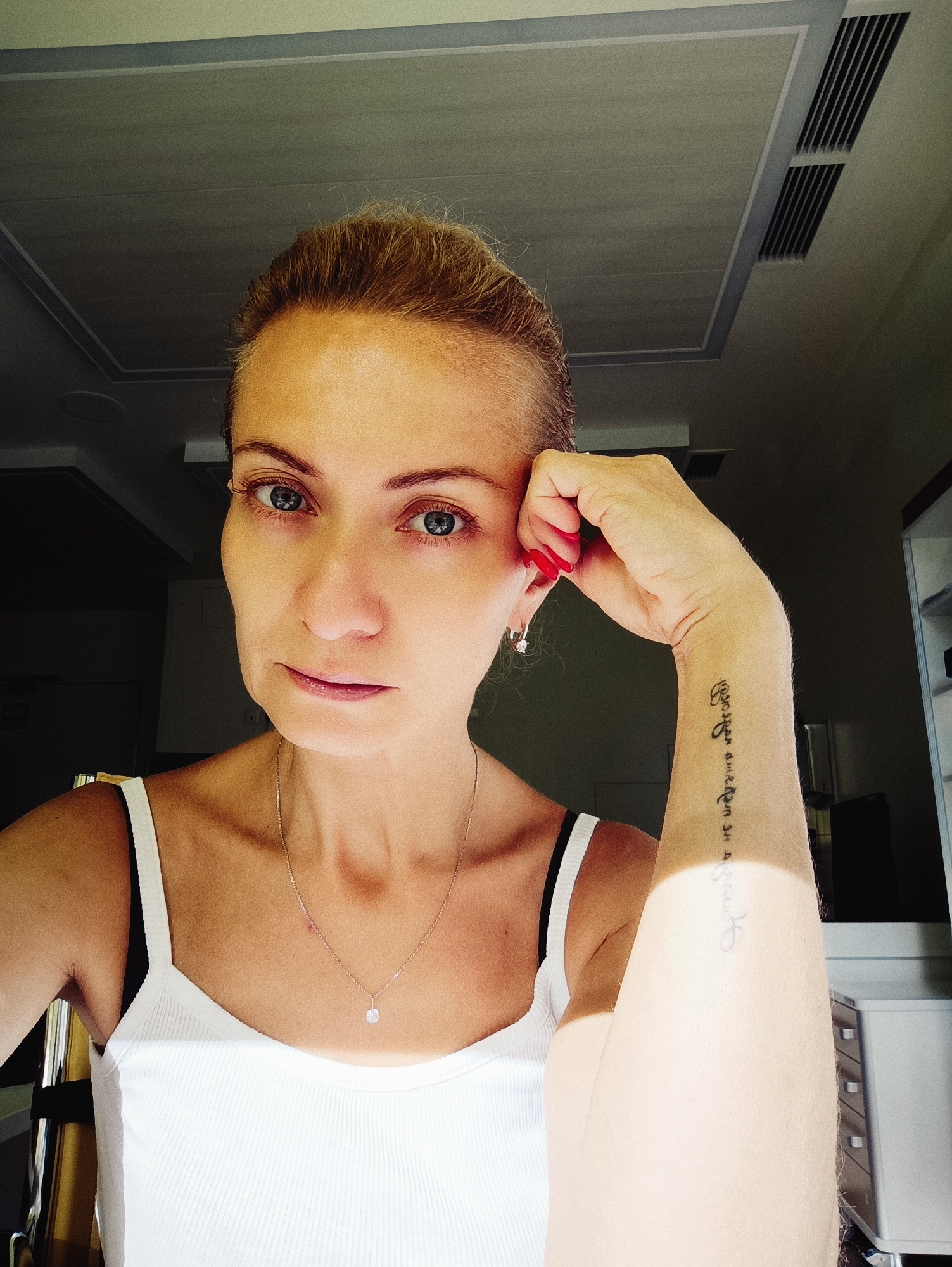A medium shot shows a woman with blonde hair pulled back, looking directly at the camera. She is wearing a white tank top and a delicate silver necklace with a small pendant. Her right arm is propped up, resting her chin on her hand, revealing a tattoo on her forearm. The woman has blue eyes and is wearing red nail polish. The background is dimly lit, showing what appears to be an indoor environment with a ceiling that has recessed lighting and ventilation. The overall lighting suggests natural light is entering from the side. There is no discernible signage or identifying markers to indicate a specific location within Moscow, Russia.
