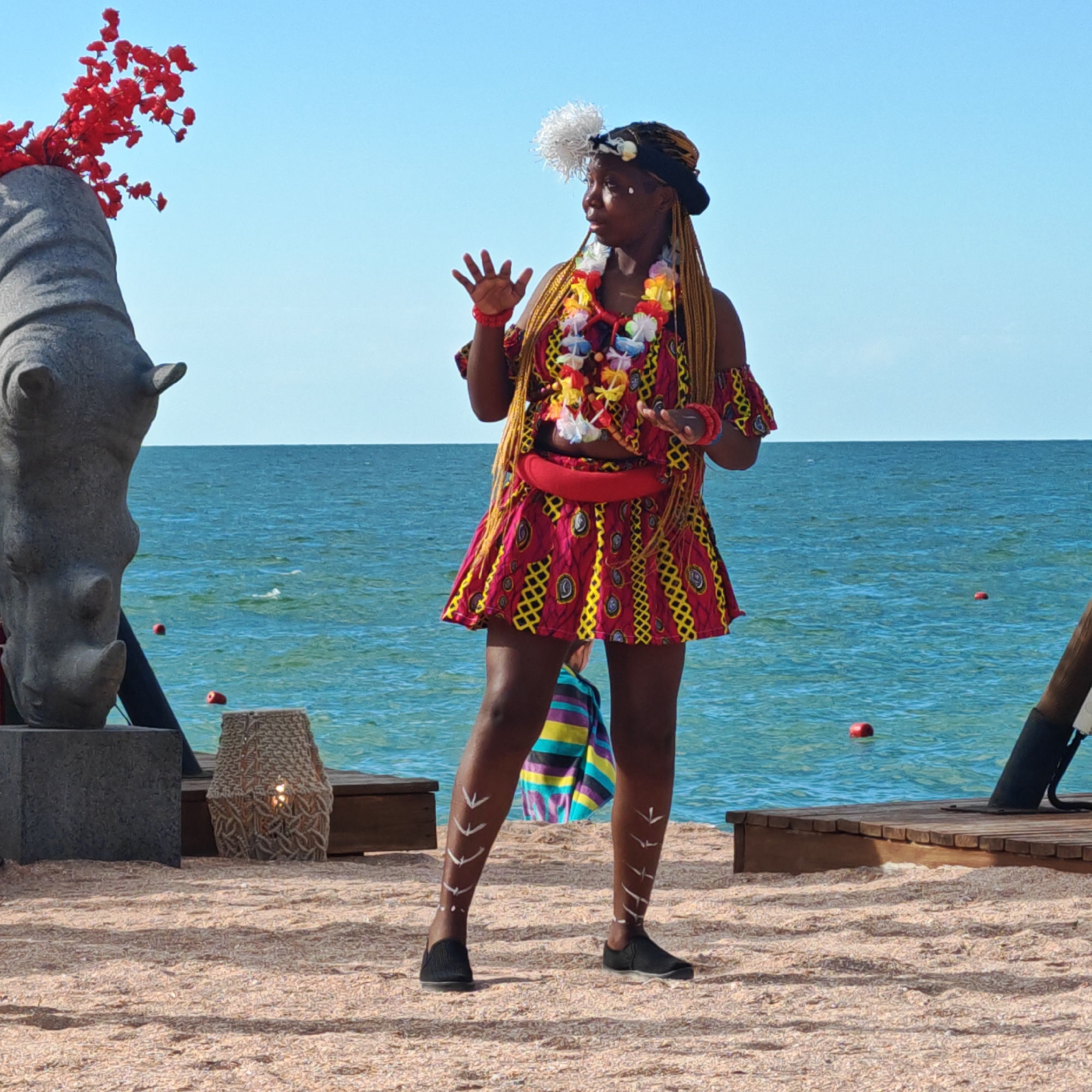A woman in vibrant, traditional African attire stands on a sandy beach, performing a dance. Her outfit consists of a colorful, patterned off-the-shoulder top and a matching short skirt, adorned with a red sash. She wears a lei of flowers around her neck and a white feathered headdress. Her arms are outstretched, and her hands are in a gesture of movement, suggesting she is mid-performance. White markings are painted on her shins. She is wearing black slip-on shoes.
Behind her, the blue sea stretches out to the horizon under a clear blue sky. To her left, a large stone sculpture of a rhinoceros head with red flowers is visible. In the background, a wooden deck is laid out on the sand, and a woven lamp with a lit candle sits on the deck. The scene suggests a cultural event or festival taking place outdoors on a sunny day. The overall atmosphere appears lively and festive. The image was taken in Shchyolkino, Ukraine. There is no visible text in the image.