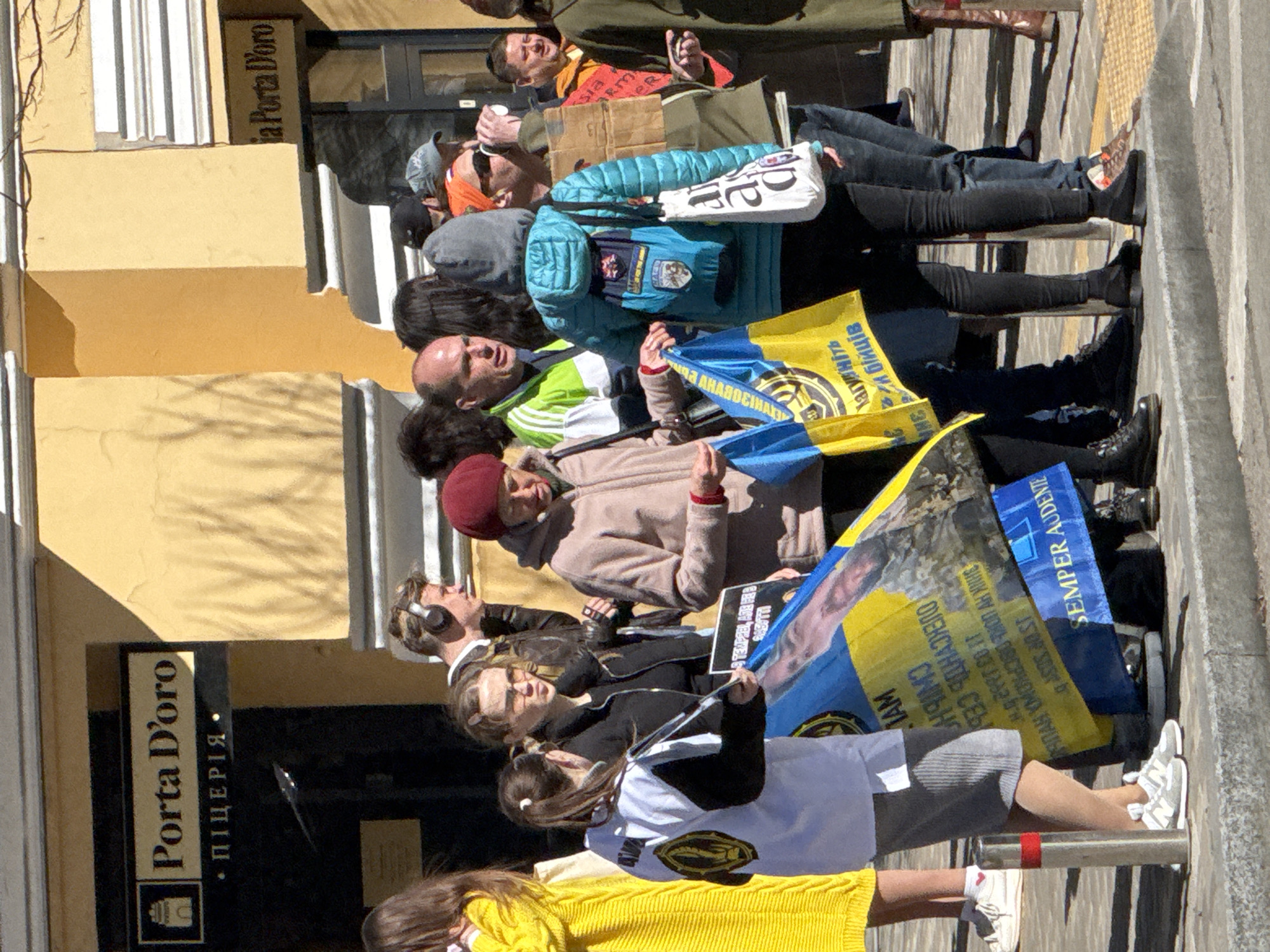 A group of people are lying on the sidewalk in Kyiv, Ukraine, in front of a building that includes a restaurant named "Porta D'oro." Some individuals are holding Ukrainian flags and signs, suggesting a protest or demonstration. The people's clothing varies, including jackets, sweaters, and athletic wear. The surrounding environment includes a building with beige-yellow walls, a doorway with a sign, and a paved sidewalk. The overall scene suggests a public gathering or demonstration taking place in a European urban setting.