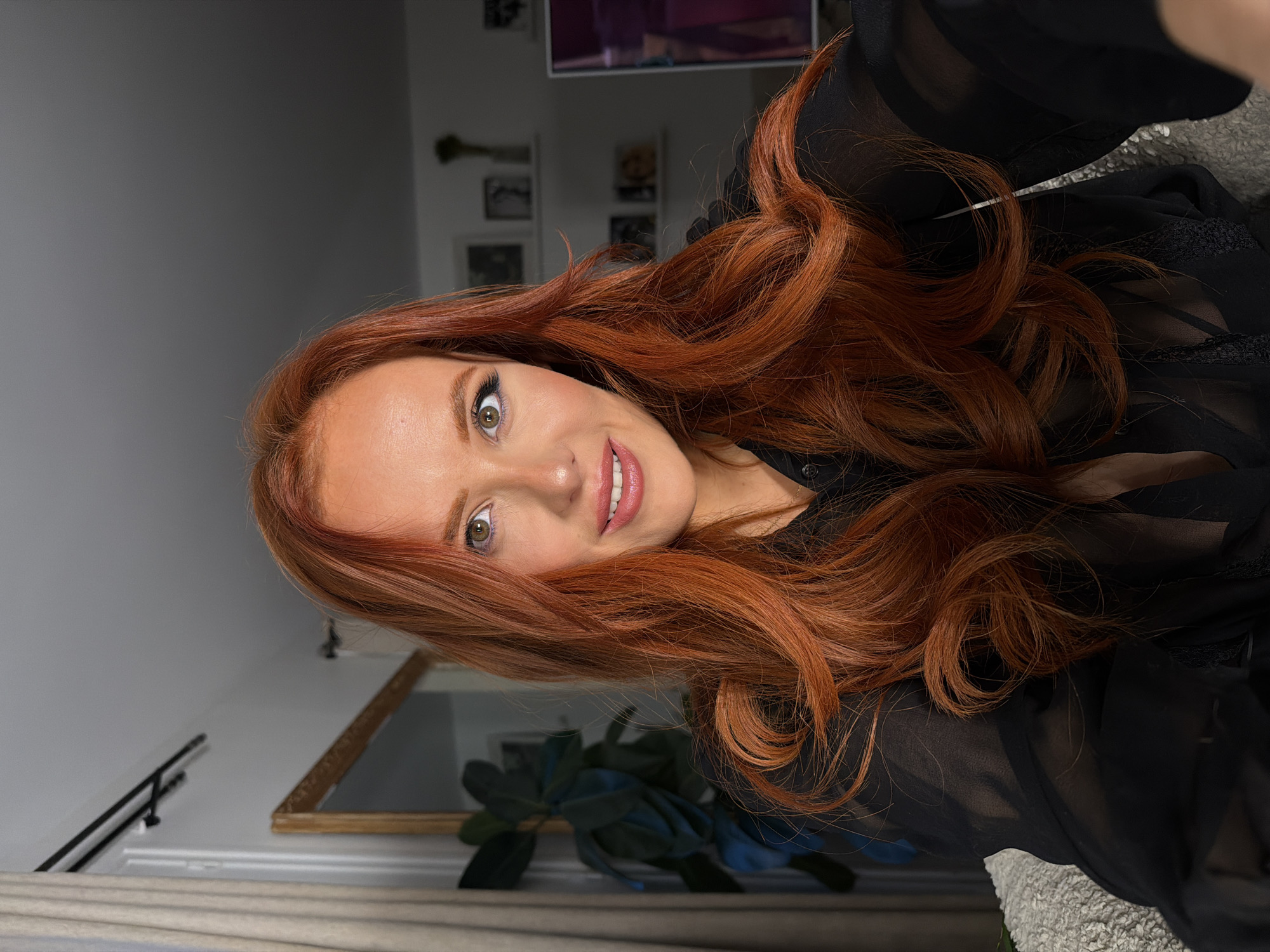 A woman with long, wavy auburn hair and light skin smiles at the camera. She wears a sheer black blouse with lace detailing. The background includes a room with a neutral-colored wall, a mirror, framed photos, and a television. The location in the city of Paris, France is not discernible from the image.