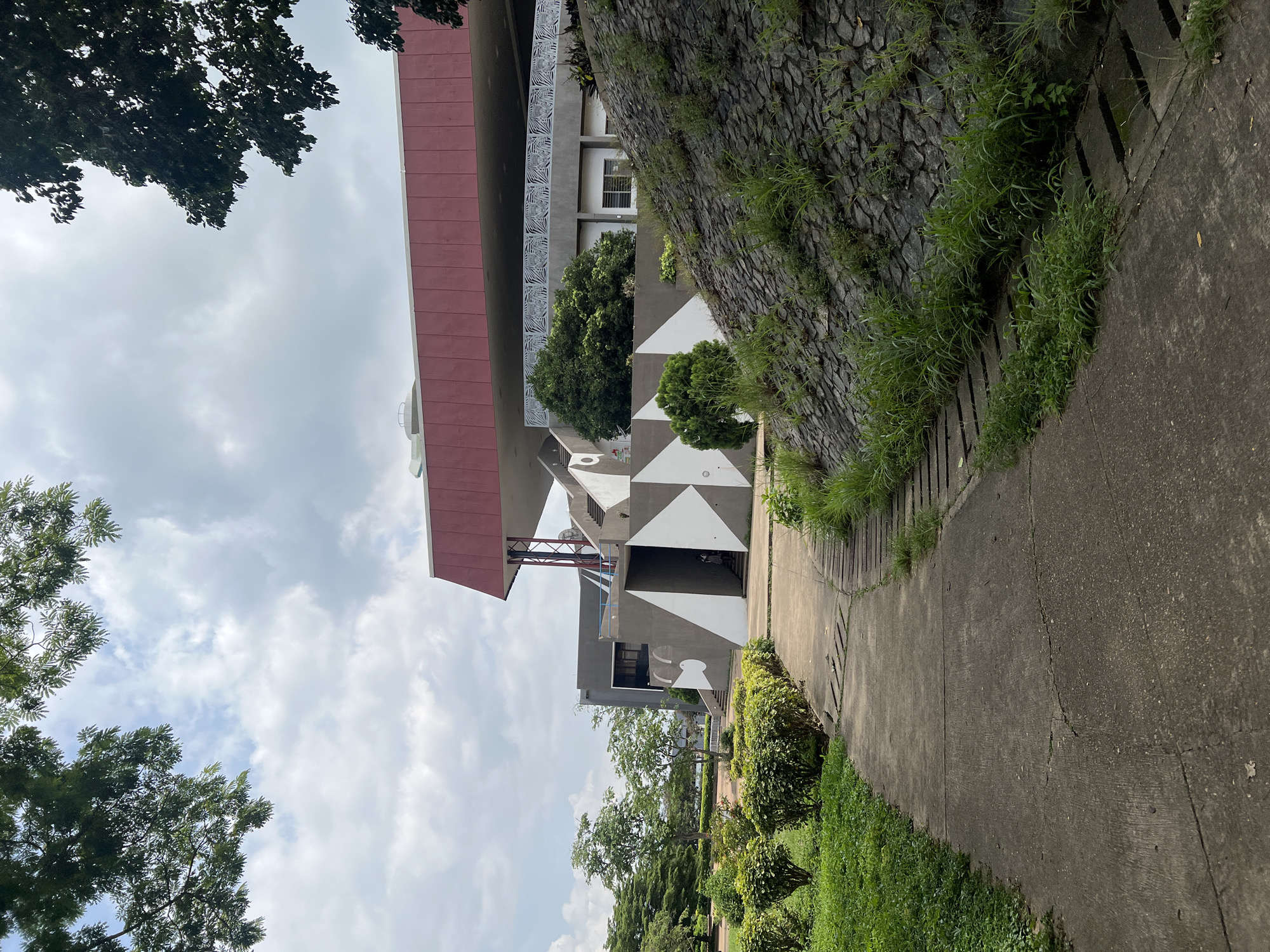 The image captures a view of the Oduduwa University Senate Building in Ile-Ife, Nigeria, under a partly cloudy sky. A concrete pathway winds through a green lawn with manicured bushes on the left. To the right, a sloping embankment is covered with stones and overgrown grass. The building features a prominent red roof, a gray facade with geometric white patterns, and visible staircases. Trees are scattered throughout the scene, with branches from a large tree framing the top of the image.