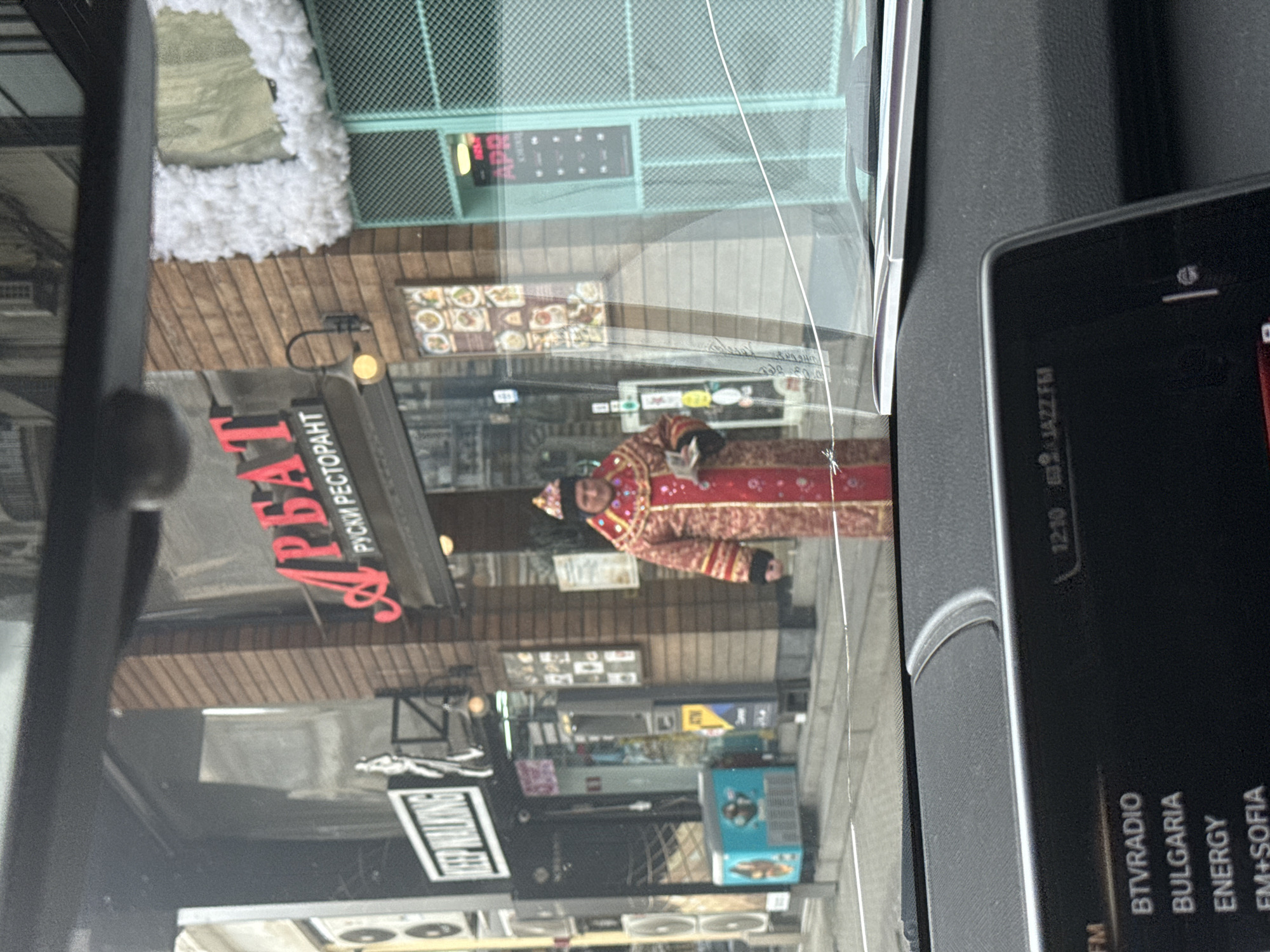 The image, taken from inside a vehicle, shows a street scene in Sofia, Bulgaria. Visible through the windshield is a man dressed in a traditional red and gold costume. To the left, the sign for the "Arbat" Russian Restaurant is prominent. Other storefronts and buildings line the street. The vehicle's dashboard is also visible, displaying radio station information including "BTVRADIO BULGARIA ENERGY FM+SOFIA" and the time "12:10 JAZZ FM". A sign reading "KEEP WALKING" is partly visible.