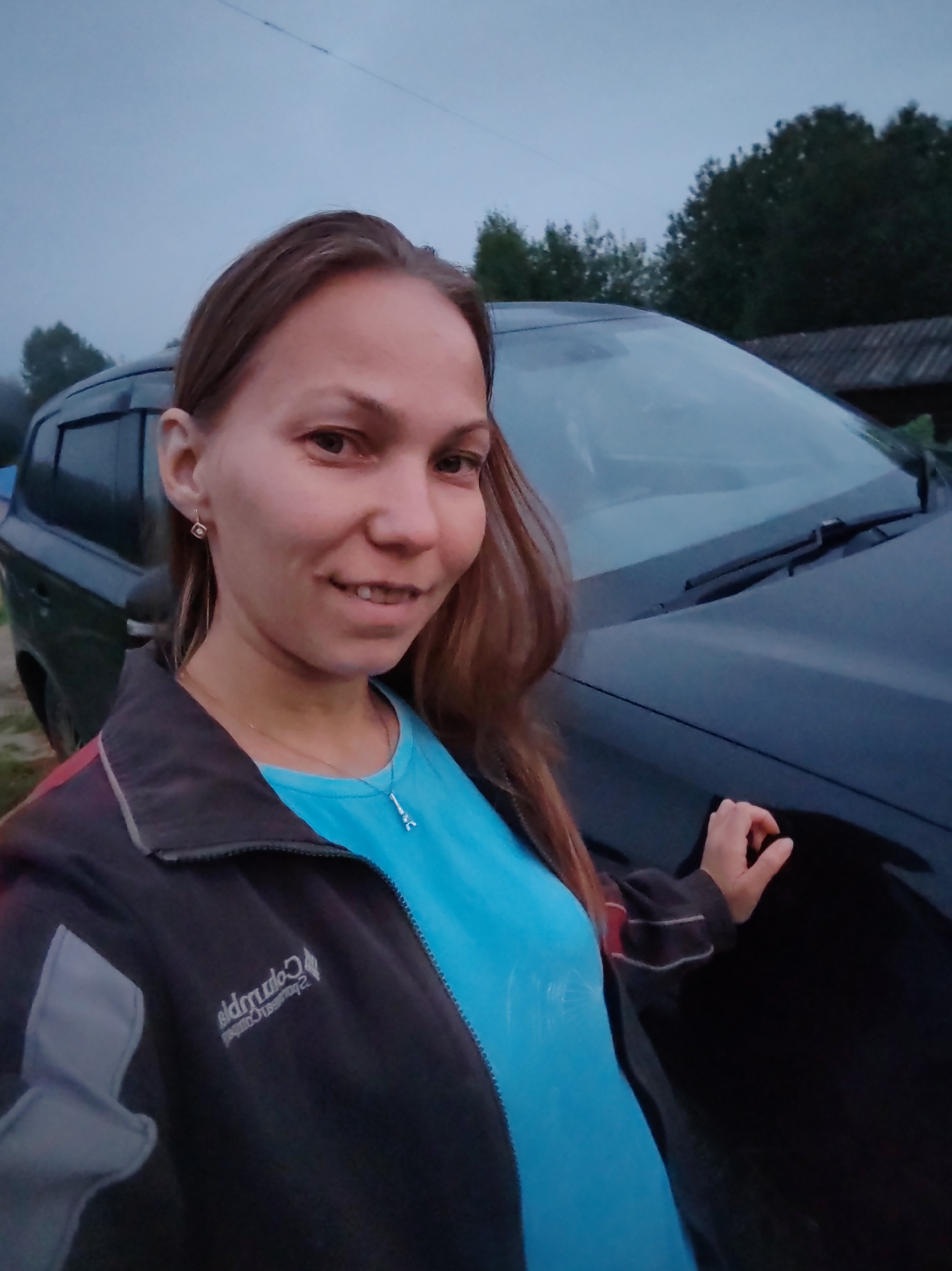 A woman with long brown hair is taking a selfie outdoors in what appears to be a rural or suburban area. She is wearing a dark jacket with gray and red accents and a bright blue t-shirt. A silver necklace with a small pendant is visible on her t-shirt. She is smiling and standing next to a dark-colored car. The background shows trees and a building with a corrugated roof, suggesting a location in a village or on the outskirts of a town. The sky is overcast. There is no visible indication that this image was taken in the city of Чухлэм, Russia.