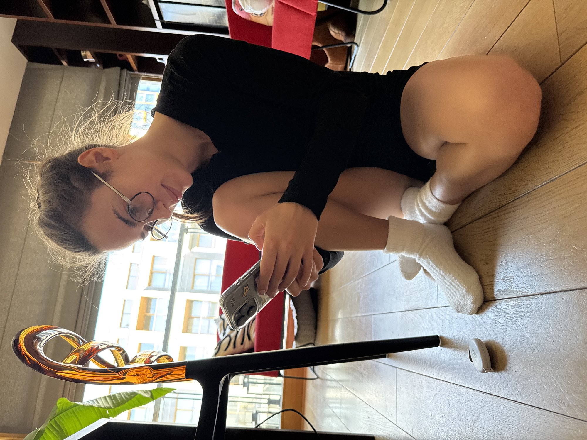 A young person is depicted indoors, squatting or crouching on a polished light wooden floor, engrossed in their smartphone. The subject, appearing female, wears a black long-sleeved top and white ribbed socks. They have light brown hair, with some flyaways, and are wearing round, metal-rimmed glasses, looking down intently at the phone held in both hands. The smartphone is enclosed in a clear case that features subtle, possibly cartoonish, designs.
The scene is set in what appears to be a modern apartment or living space in Warsaw, Poland. A large window in the background provides ample natural light and a view of external urban buildings, suggesting a daytime setting. To the left, a distinct amber-colored, pretzel-shaped glass art piece or decorative object rests on a black table or stand, beneath which a vibrant green plant leaf is partially visible. Further back, part of a red chair and an object with a tiger-stripe pattern are visible. A small, white, elliptical device or object lies on the wooden floor near the person's feet. The overall impression is one of casual focus within a brightly lit, contemporary interior. No readable text is visible in the image.