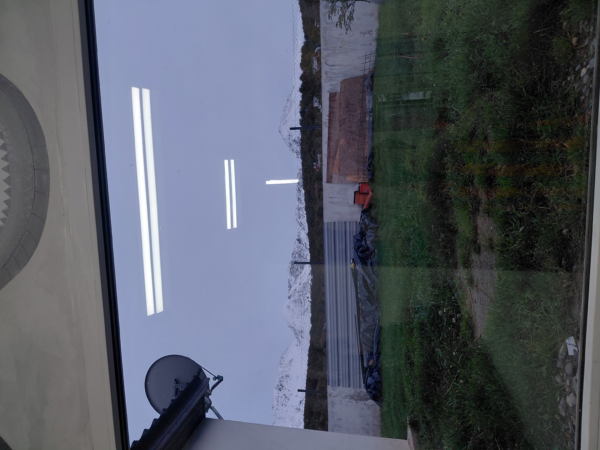 The image shows a view from inside a building, looking out a window. The location is Ushuaia, Argentina, as evidenced by the distant view of the snow-covered Andes mountains, a characteristic feature of the landscape around the city. The foreground shows a grassy area and a building exterior with a satellite dish attached to the roof. Construction materials, including corrugated metal sheets and possibly wooden boards, are visible near a concrete wall in the middle ground. The interior reflections on the window show what appears to be fluorescent light fixtures, providing a contrast to the outdoor scenery. The absence of people suggests the scene is uninhabited at the time of capture.