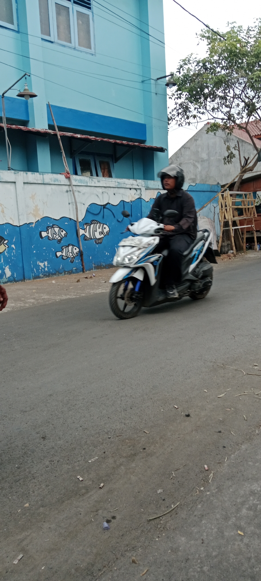 The image shows a person riding a white and blue scooter on a paved road in front of a blue building with a mural of fish on its lower wall. The building has windows and a blue band running across its facade. The road surface is gray asphalt with some debris. To the right of the building, there is a grey concrete wall and a tree with green foliage. The scooter rider is wearing a black long-sleeved shirt, black pants, and a black helmet with a clear visor. The scooter is a typical automatic scooter with its front wheel in motion. The lighting suggests it is daytime, possibly overcast. The overall scene depicts a street in an urban or semi-urban area.