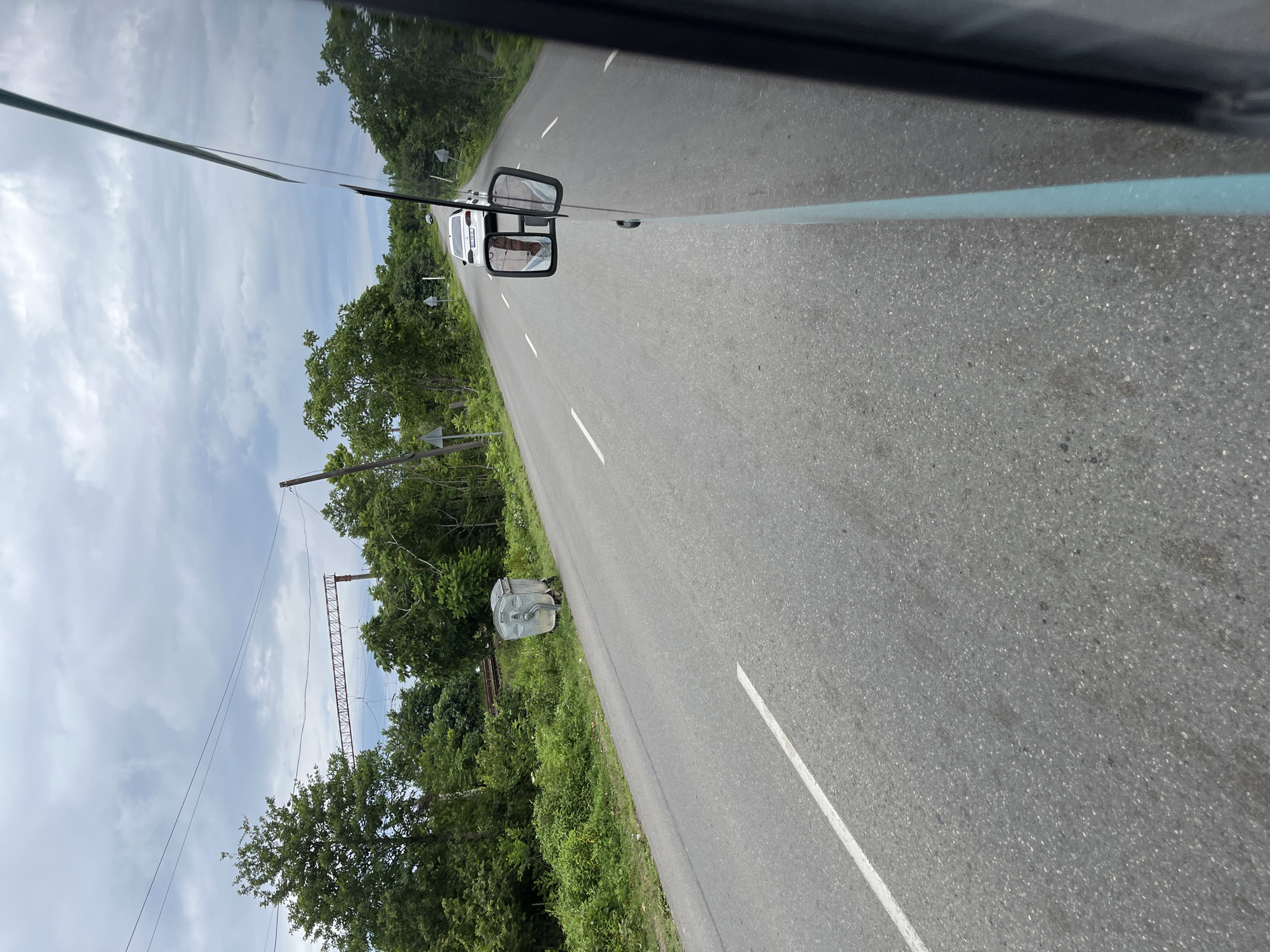 The image captures a view from a vehicle traveling on a paved road in Ureki, Georgia. The road is lined with lush green trees and vegetation on both sides. In the foreground, the right side of the image shows the exterior of the vehicle, including a side-view mirror reflecting the interior and a person. The road has a white line marking the lane. Further down the road, a white car is visible. In the distance, there are power lines and a rusty metal structure that appears to be part of an overhead railway or industrial equipment. On the left side of the road, near a clump of trees, a large metal trash bin on wheels is present. The sky is overcast with gray clouds. The scene suggests a rural or suburban setting within Ureki.