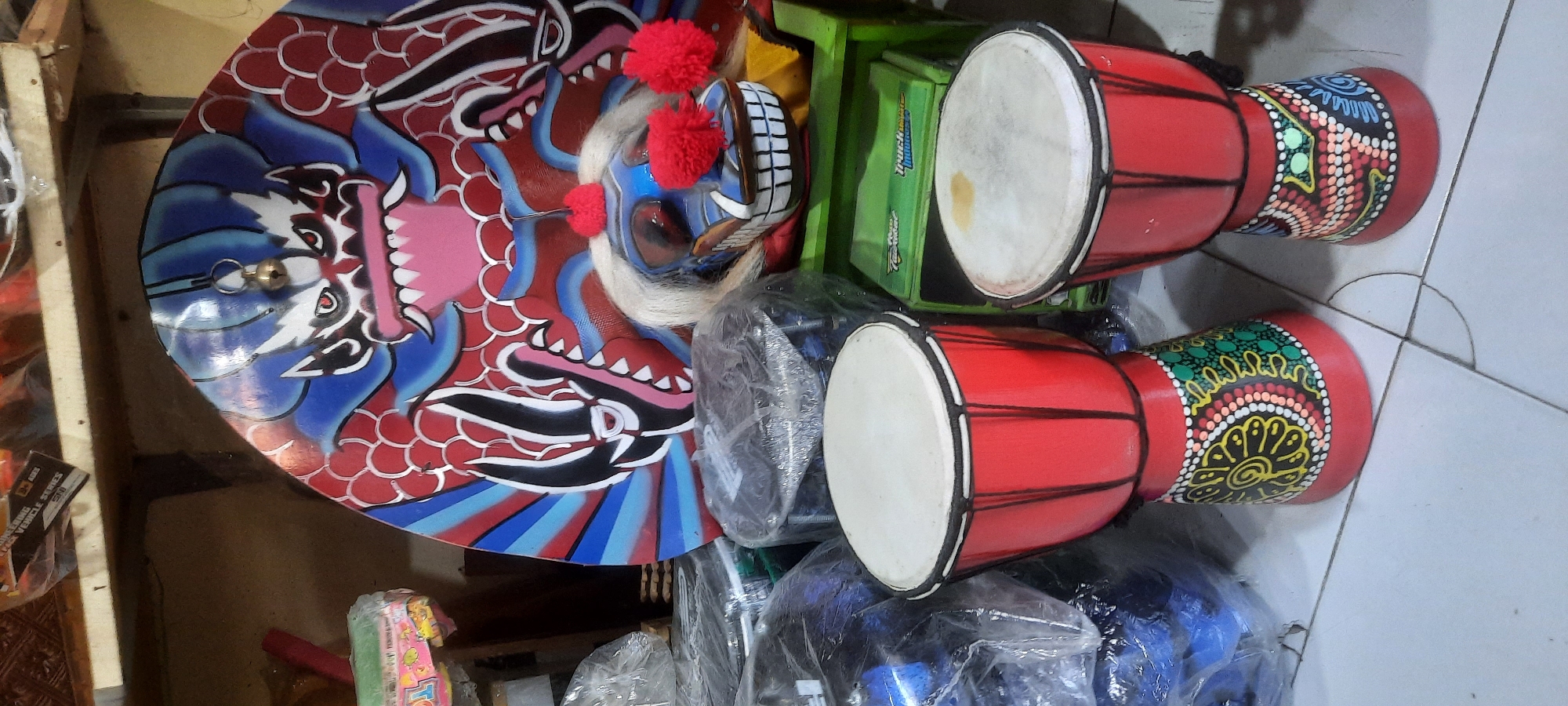In Blitar, Indonesia, a close-up shot captures a collection of handcrafted items, likely for sale or display in a market or shop. Dominating the foreground are two red djembe drums, each adorned with intricate, colorful dot patterns around their bases. The drum on the left is slightly taller and features a mandala-like design with yellow, green, pink, and white dots, while the drum on the right has a more abstract pattern with similar colors. Behind the drums, a large, vibrant mask with a fierce, dragon-like face painted in red, blue, and black is propped up. This mask has a prominent golden bell attached to its forehead. To the right of the mask, a smaller, blue and white mask with a similar fierce expression and decorated with fluffy red pom-poms rests on a green shelf. The background reveals stacks of items wrapped in plastic, possibly musical instruments or other merchandise, and a glimpse of shelving with various objects. The scene is set against a tiled floor, indicating an indoor setting.