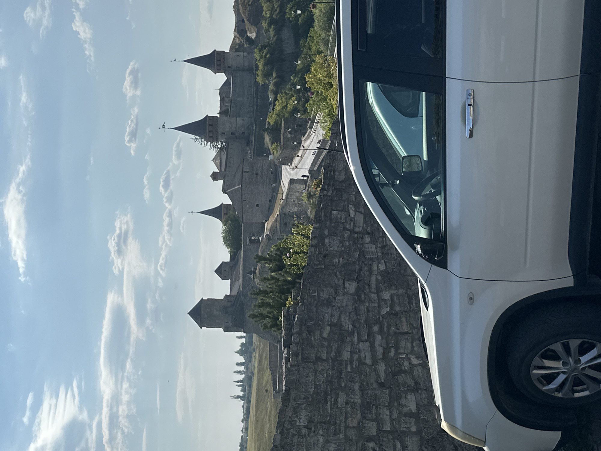 In the foreground, the right side of a white SUV is visible, parked on a dark surface. The vehicle's front right wheel and fender are in view, along with its side door and door handle. Behind the SUV, a stone wall runs horizontally across the lower portion of the frame. Beyond the wall, a panoramic view unfolds of the Kamianets-Podilskyi Castle, a historic fortress situated in Kamianets-Podilskyi, Ukraine. The castle, constructed of stone with multiple towers and turrets, is perched on a rocky outcrop surrounded by greenery. A pathway or road leads up to the castle, with lampposts lining its edges. The sky above is a pale blue with scattered white clouds, and a few trees are visible on the distant horizon.