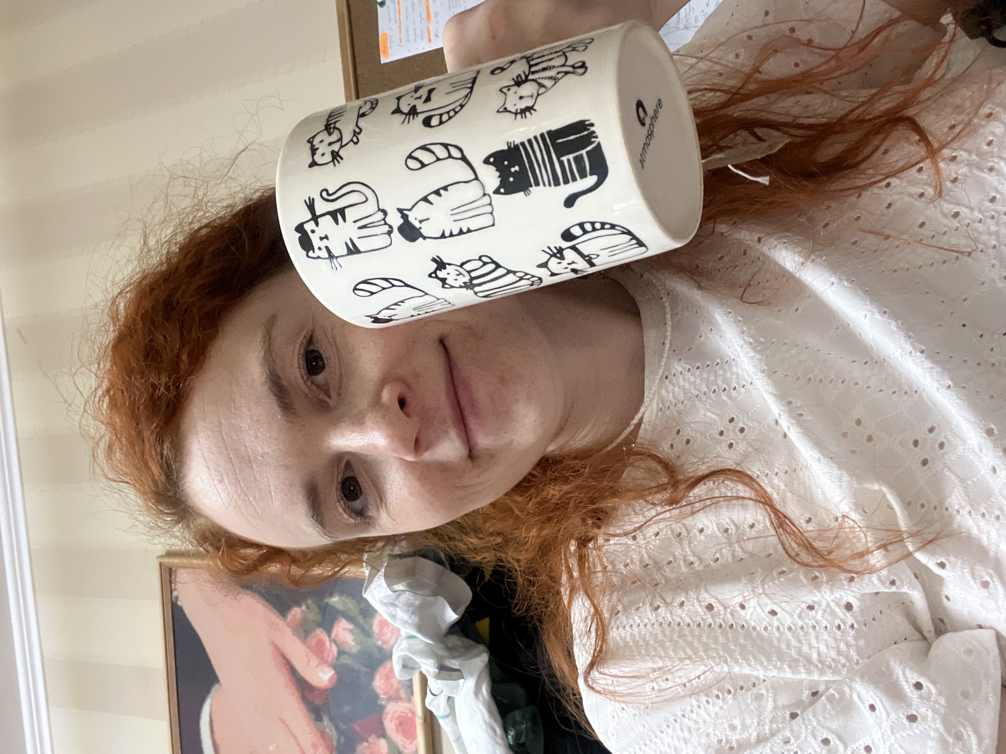 A Caucasian woman with reddish-brown hair holds a white mug with black cat illustrations in front of her face. The mug has the word "atmosphere" printed on its side. The woman is wearing a white, embroidered shirt. Behind her, there is a framed painting of roses, and a bulletin board is partially visible at the top. The location of Chaikovsky, Russia, cannot be verified based on the image.