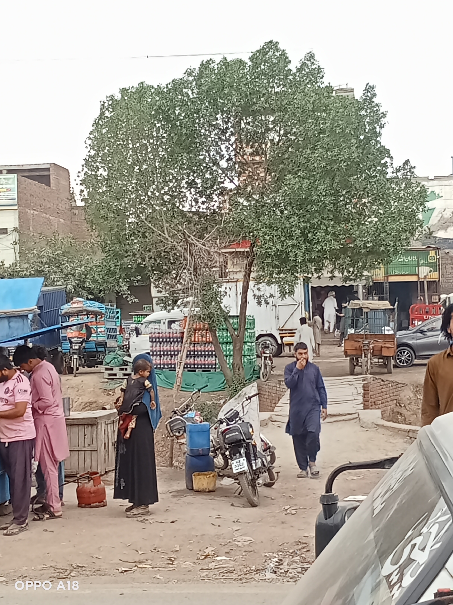 The image shows a street scene in what appears to be an urban area. A large tree dominates the background. A group of men in pink shirts stand on the left. A woman in a long black dress holds a child. A man in a dark blue outfit walks on a bricked walkway, talking on a cell phone. A black motorcycle is parked nearby. A truck with soda cases in the bed is behind the tree. In the distance, people are walking into a store. The ground is dirt. Part of a vehicle is visible on the right side of the image. The bottom left corner shows the text "OPPO A18."