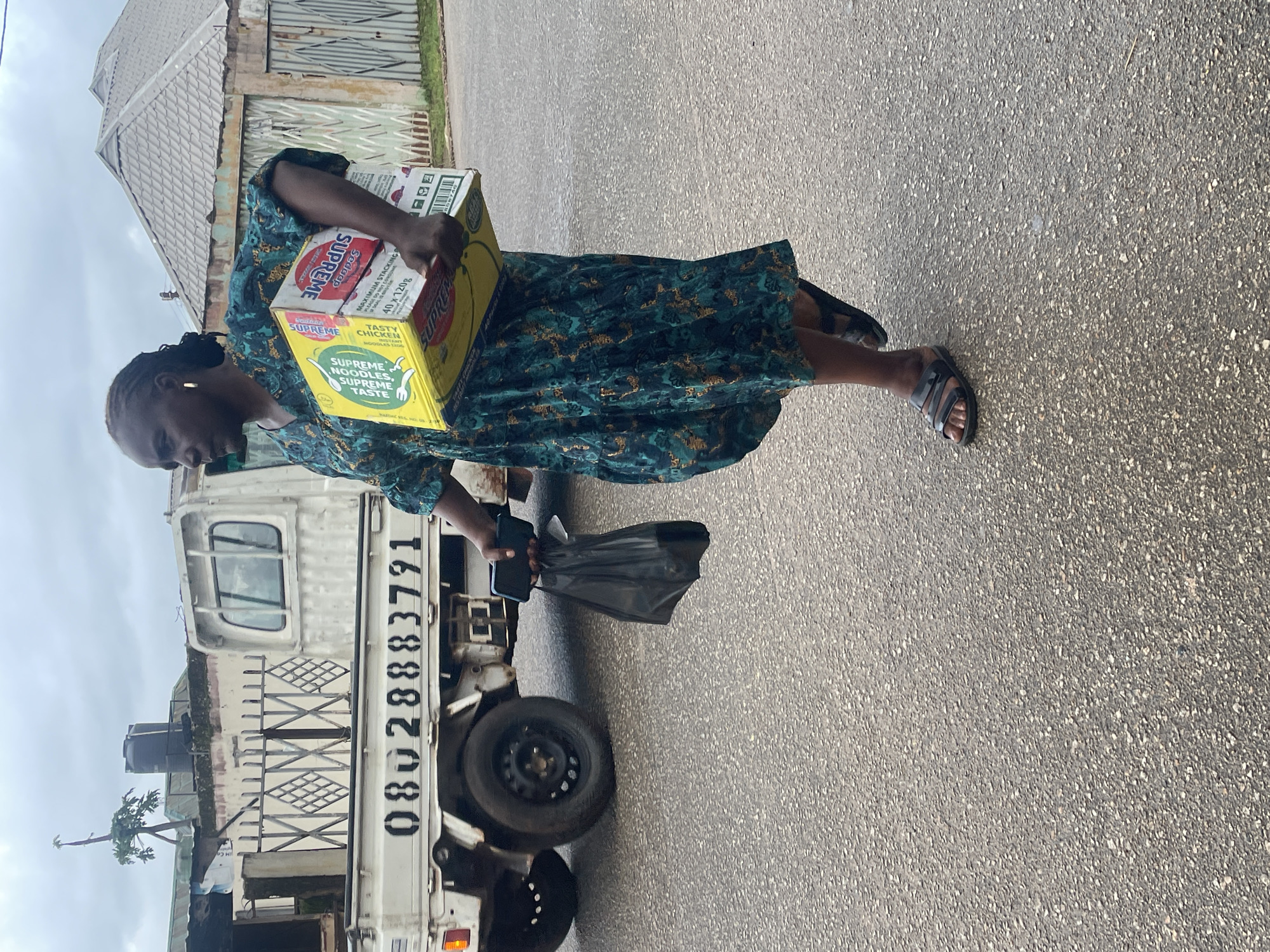 An overcast day in Jos, Nigeria, is captured in this medium shot of a woman walking on a paved road. She is in motion, her body angled to the right, her gaze directed forward. The woman, wearing a dark green dress with a floral print and black sandals, carries two boxes in her right arm and a black plastic bag in her left hand. The boxes, stacked atop each other, are branded with "SUPREME" in red lettering. One box is yellow and depicts a cartoon chicken, while the other is white and red with text indicating "MAXIMUM STACKING". A silver-colored cell phone is visible in her left hand, held alongside the plastic bag.
Behind the woman, a white flatbed truck is parked. Its side displays a black phone number: "08028883791". The truck is adorned with a metal railing along its bed, and a black water tank sits atop a structure behind it. The background reveals a residential area with a gray-roofed house and a corrugated metal fence. The road surface is asphalt, speckled with small stones. The overall lighting suggests diffused daylight.