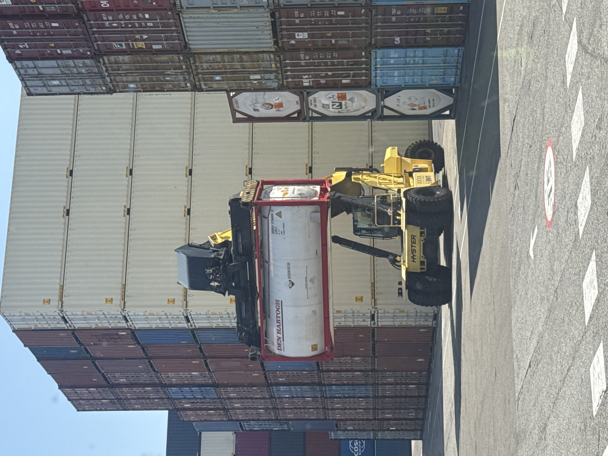 Here's a forensic description of the image:
The image depicts a scene at what appears to be a container terminal, based on the presence of stacked shipping containers. A yellow Hyster reach stacker, a type of heavy-duty forklift, is lifting a red-framed tank container. The tank has "DEN HARTOGH" written on it. The side of the tank container is labeled "DHOU 215809 9 22K2".
The containers are stacked in rows and columns. Various colors of containers are visible, including blue, red, white, and gray. Some of the containers feature hazard symbols, suggesting they might contain specific types of cargo.
The ground surface consists of paved concrete with painted road markings, including speed limits and directional indicators.
Given the context of container shipping and the company name "Den Hartogh", the likely location is Botlek Rotterdam, Netherlands, a significant port and industrial area known for its chemical and logistics facilities.