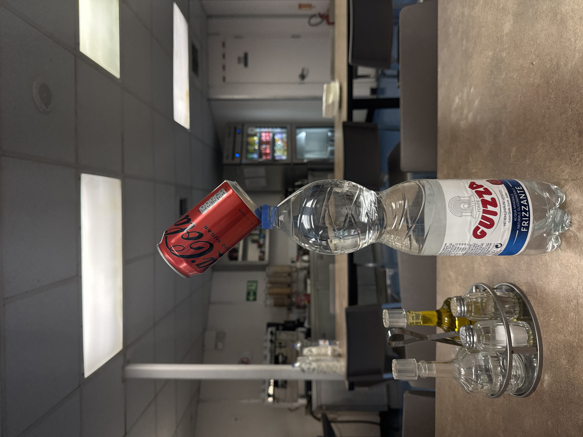 An overhead view captures a precarious balancing act on a tabletop. A red Coca-Cola Zero Sugar can is tilted and resting on the cap of a clear plastic water bottle. The water bottle, filled with clear liquid, is itself balanced on top of another identical water bottle, creating a vertical stack. In the foreground, a condiment caddy holds a glass cruet, a salt shaker, a pepper shaker, and a small bottle of olive oil. Behind the bottles, blurred figures are seated at tables in a brightly lit room with white acoustic ceiling tiles and fluorescent lighting. The background shows a kitchen area with various appliances and shelving. The scene appears to be set in a cafeteria or communal eating area.