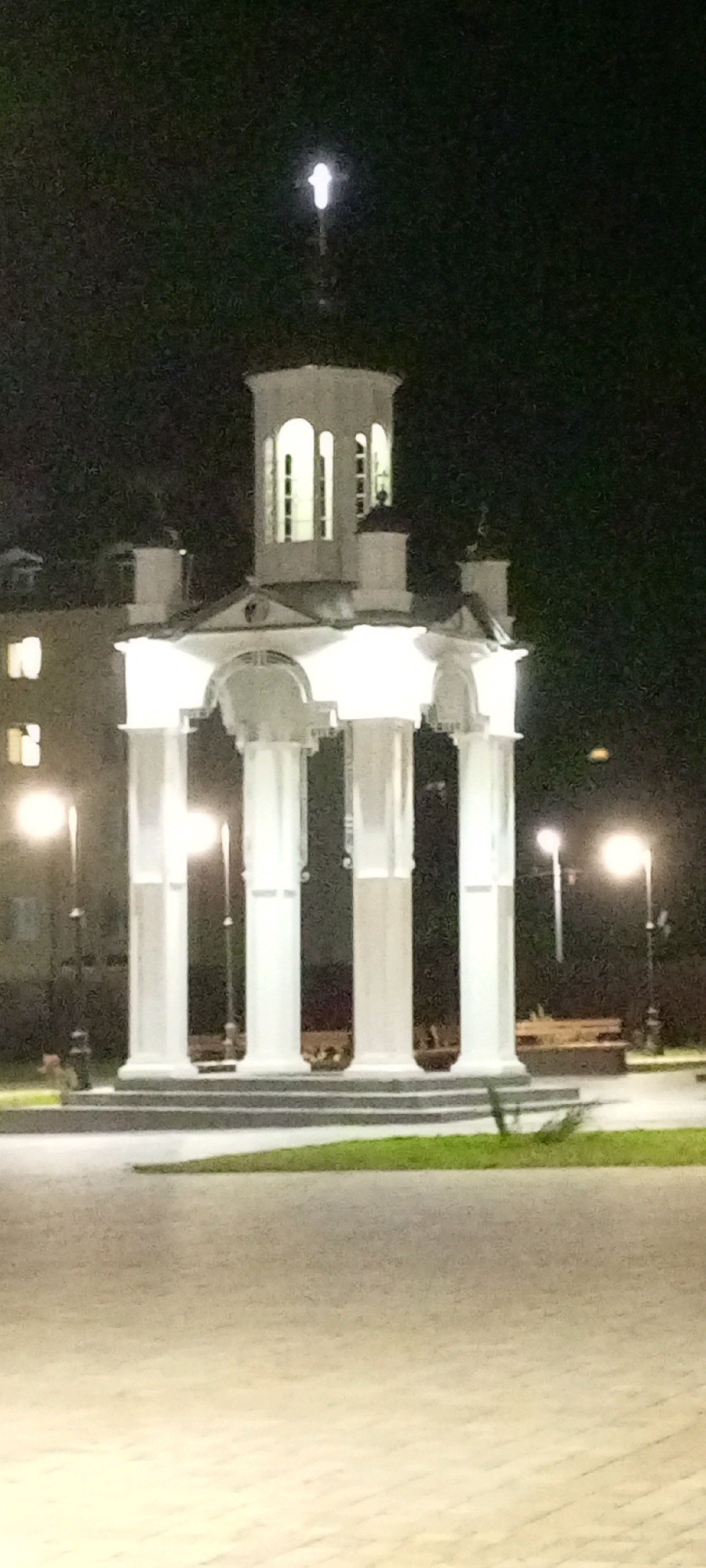 The image captures a white, multi-tiered monument with classical columns at its base, illuminated at night. The monument has a circular upper section with arched windows and a finial at its peak. The scene is set against a dark, possibly night sky, with lights from nearby buildings visible to the left, showing illuminated windows. Streetlights are present on the right side of the monument, casting a warm glow. The ground in front of the monument is paved with light-colored interlocking stones, bordered by a strip of green grass. Benches are visible in the background, suggesting a public space or park. The overall impression is a well-lit architectural landmark in a tranquil urban setting. The monument is located in Kirsanov, Russia.
