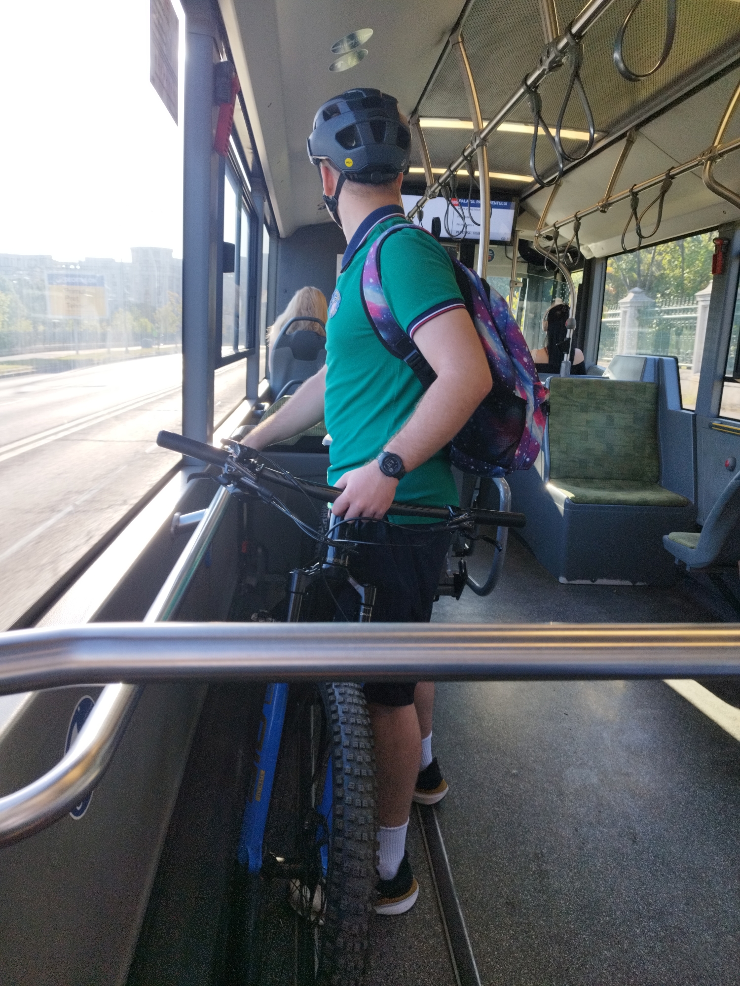 The image depicts the interior of a public transport bus, likely in Bucharest, Romania. A person wearing a green polo shirt, black shorts, white socks, and athletic shoes is standing and holding the handlebars of a blue bicycle. They are also wearing a black helmet and a backpack with a galaxy print. The bus appears to be in motion, with the exterior visible through the windows. The seating inside the bus is a muted green color. Other passengers are partially visible in the background, including a person with blonde hair. The lighting suggests it is daytime, possibly late afternoon or early morning, given the angle of the sun. The bus interior has overhead handrails for standing passengers.