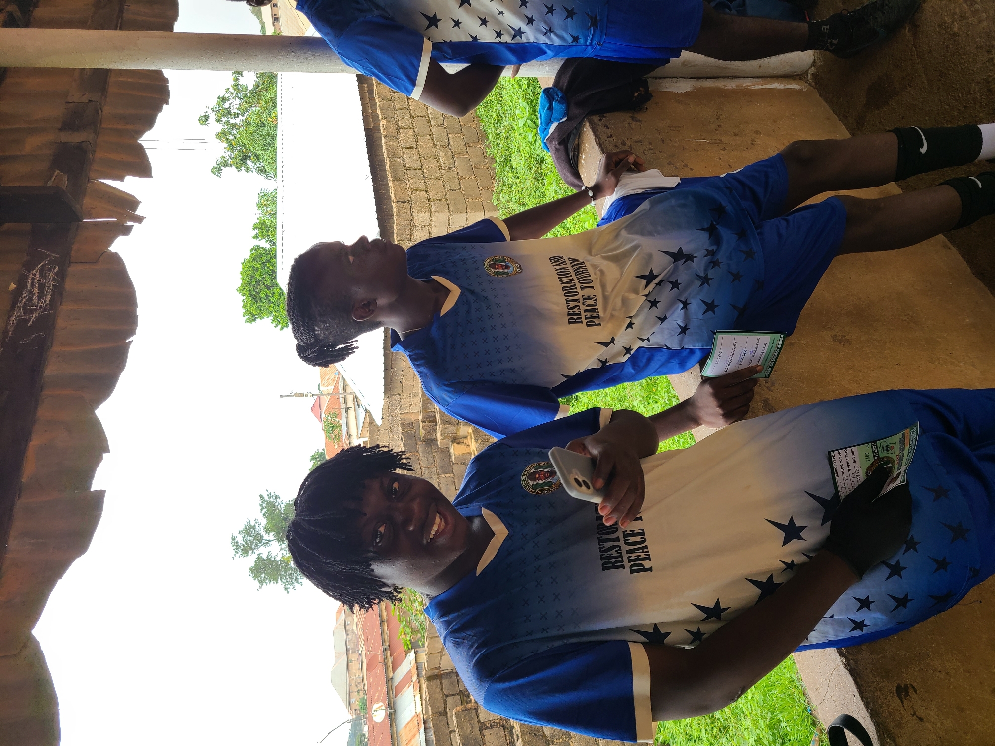Two young men are sitting next to each other, likely outdoors, wearing matching blue and white athletic shirts with the words "RESTORATION AND PEACE TOURNAMENT." The first man is holding a cell phone and smiling at the camera. The second man is looking to the side. The surrounding environment includes buildings, greenery, and a partial view of a roof. A third person wearing a matching shirt is partially visible behind them. Both individuals are holding documents.