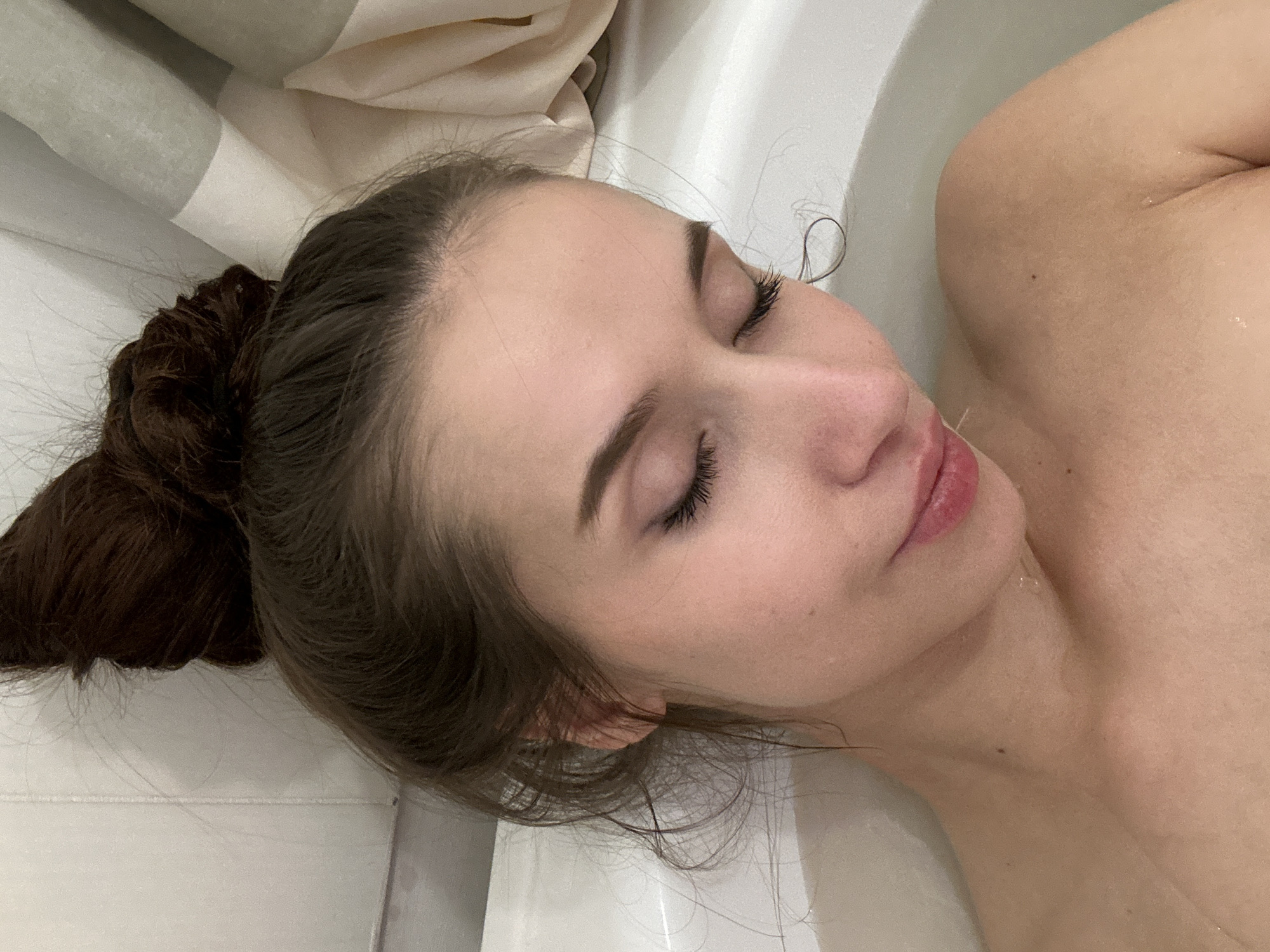 A woman is reclining in a bathtub. Her eyes are closed. Her hair is pulled back in a bun. She is partially submerged in water. A shower curtain is visible in the background. The location is unspecified and there is no indication the scene is in Yekaterinburg, Russia.