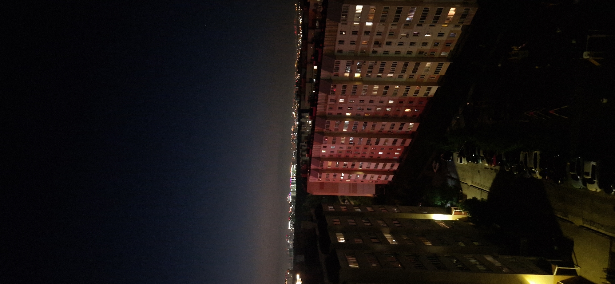 This is an aerial, nighttime photograph of a residential area in Krasnoobsk, Russia. The foreground features two tall, multi-story apartment buildings. The building on the left is largely dark, with a few lit windows. The building on the right is illuminated with many lights from within the apartments, casting a reddish hue on its facade. A street runs alongside the buildings, with a row of parked cars visible. Beyond the apartment buildings, the cityscape stretches into the distance, with a hazy horizon dotted with numerous lights from other buildings and infrastructure, suggesting a larger urban area. The sky is dark, with the faint glow of distant lights visible. The overall scene depicts a typical urban night in a Russian city.