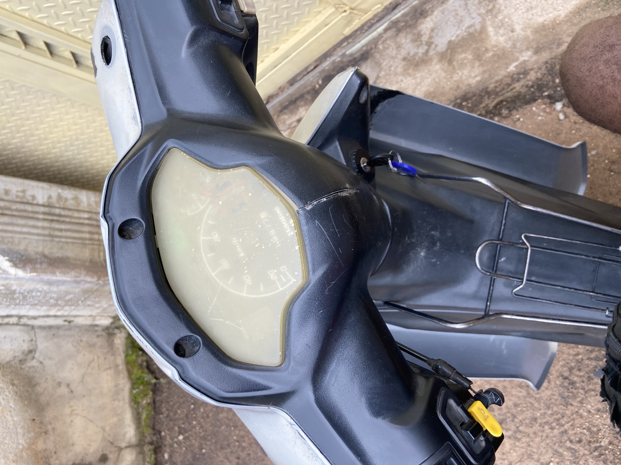 In Mararaba, Nigeria, a close-up shot focuses on the handlebars and instrument cluster of a black and silver motorcycle. The instrument cluster, with its light yellow, scratched display, shows markings for speed in km/h up to 80, and a partially visible odometer reading of "133776". Below the cluster, a key with a blue fob is inserted into the ignition. To the left, a yellow lever is visible on the handlebar. The background is blurred, showing a textured concrete wall and a yellow corrugated metal surface. The overall scene suggests an outdoor setting in a somewhat weathered environment.