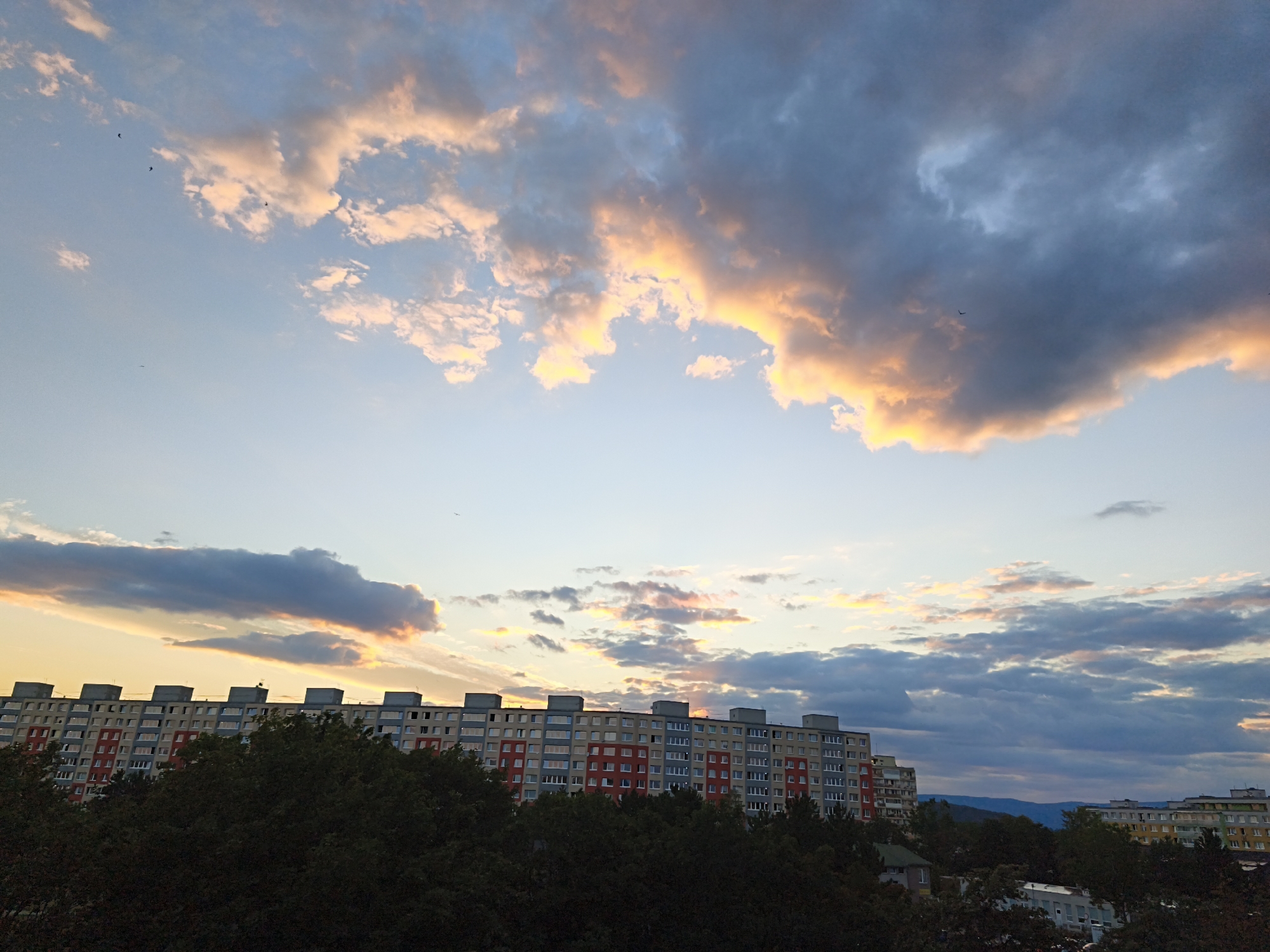 A wide-angle, high-angle shot captures a pastel sky with illuminated clouds during sunset. The sky transitions from a pale blue near the horizon to deeper blues and grays higher up, with touches of peach and yellow highlighting the edges of the clouds. Several small, dark birds are visible in flight. In the foreground, a long, multi-story apartment building, painted in sections of red, gray, and beige, stretches across the frame. The building has numerous windows and a row of distinct rectangular structures on its roof. Dense, dark green foliage, likely trees, obscures the lower portion of the building and extends to the bottom of the image. In the distance, the outline of rolling hills can be seen against the horizon, and another apartment building with a yellow facade is partially visible on the right. This scene is consistent with the "Severní sídliště" (North Housing Estate) in the city of Most, Czechia, characterized by its panel housing blocks and surrounding greenery, often experiencing dramatic sunsets due to its location.