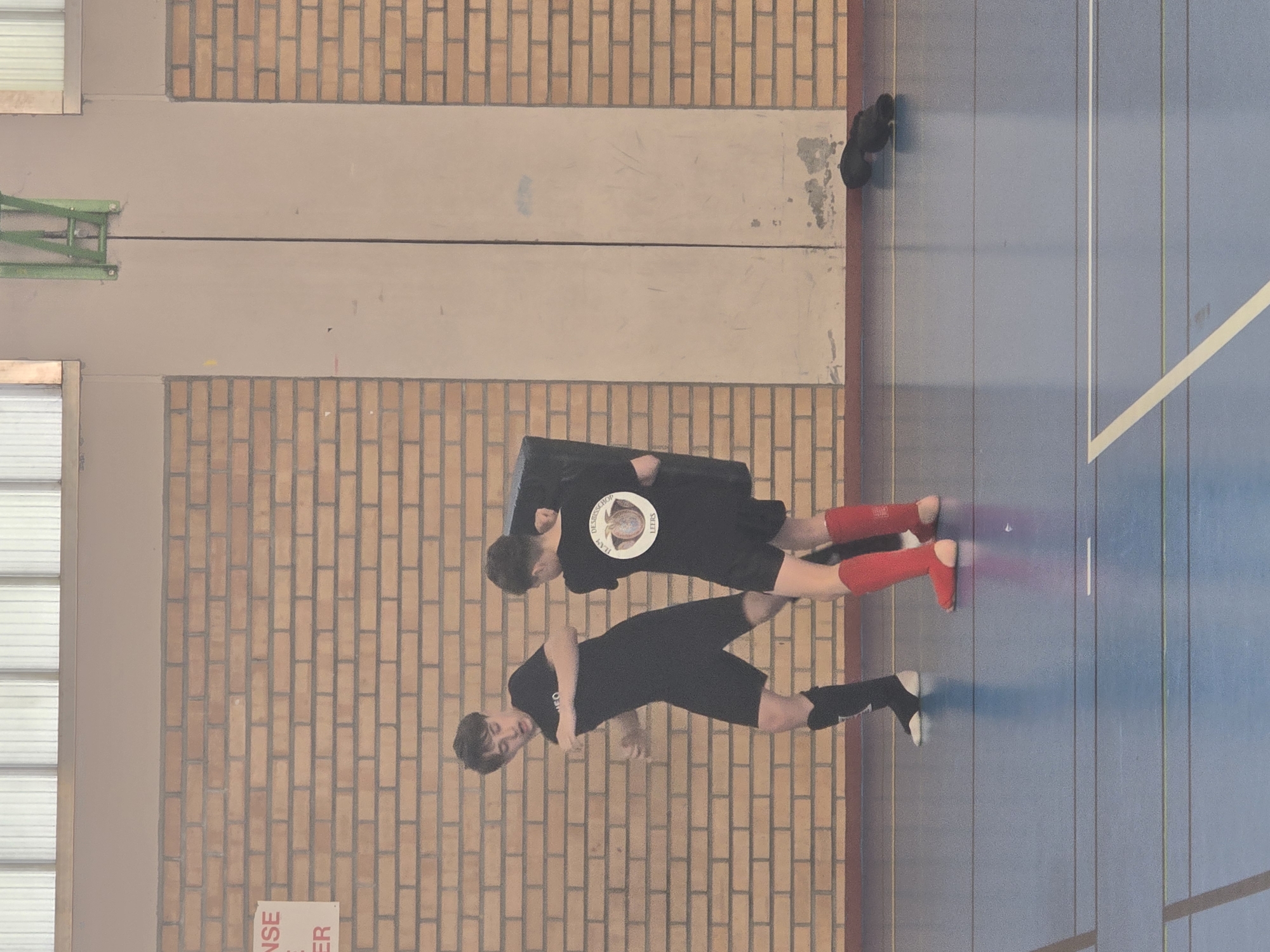 Two individuals are positioned on a blue indoor court with white lines. One person is wearing black shorts, a black shirt, and black and white shoes. The other is wearing black shorts, a black shirt with a circular logo, and red socks. They are indoors, possibly in a gymnasium, as evidenced by the brick wall, the basketball hoop, and the presence of athletic shoes placed away from the main activity. The location can be inferred as Leers, France, given the information available in the image.