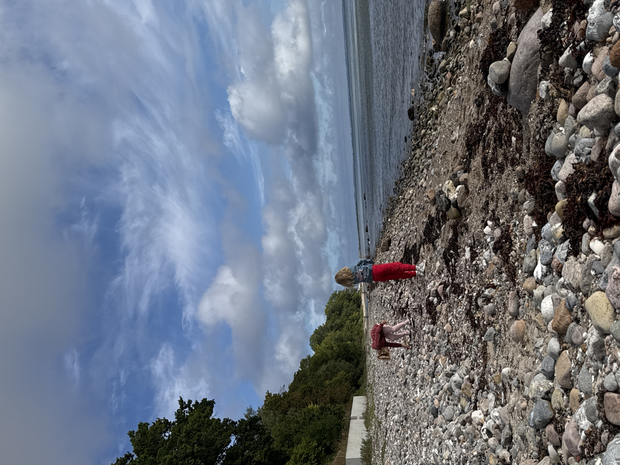 Two young girls are on a rocky beach with the sea to the right and a dense line of trees and a concrete barrier to the left. The girl on the left is bending down, appearing to examine something on the ground, wearing a pink top and light pink pants. The girl on the right stands upright, facing away from the camera, wearing a denim jacket with a design on the back and red pants. The sky is a mix of blue with scattered white and gray clouds. The tide is out, revealing a wide expanse of pebbles and seaweed on the beach.