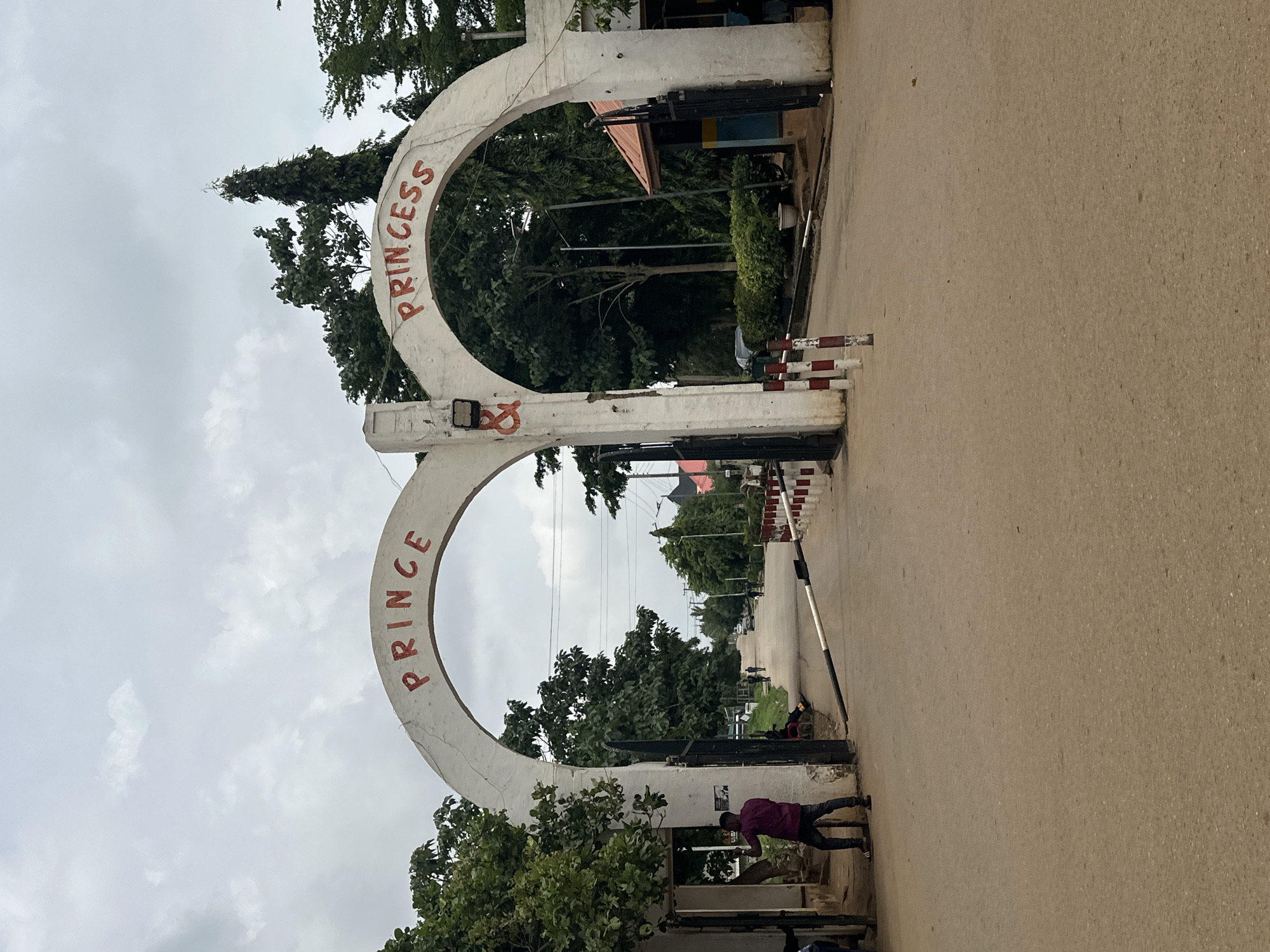 The image shows the entrance to "PRINCE PRINCESS" in Abuja, Nigeria. A large, white archway spans a paved road, with the words "PRINCE" and "PRINCESS" painted in red on either side. The archway has black metal gates on both sides, which are open.
On the left side of the archway, a man in a purple shirt and dark pants stands near the gate, holding a phone. He appears to be looking at the phone and possibly taking a photo or video. A portion of a building or wall is visible behind him.
The road continues into the distance, lined with trees on both sides. Several other people are visible in the background, walking along the road. A red and white striped barrier is partially lowered across the road in the foreground, suggesting it is a controlled entry point.
The sky is overcast and appears to be cloudy. The overall scene depicts a daytime view of an entrance to a facility or compound.