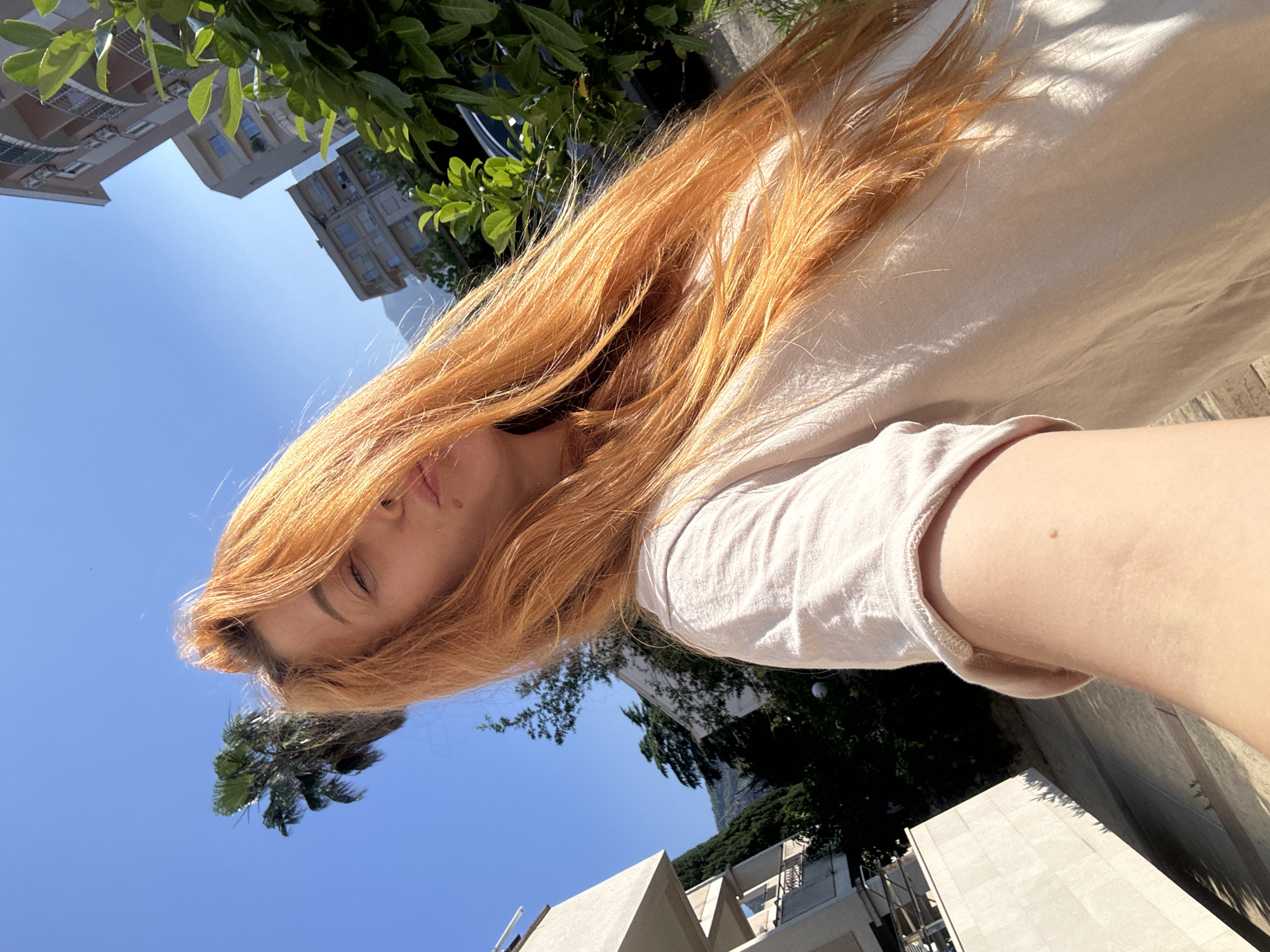 A young woman with long, reddish-blonde hair is taking a selfie outdoors on a sunny day. She is wearing a light-colored t-shirt. Her hair is mostly in front of her face, obscuring it. She is puckering her lips. In the background, there are buildings, trees, and a palm tree, suggesting an urban environment. The scene appears to be in Budva, Montenegro, as indicated by the architecture and vegetation.