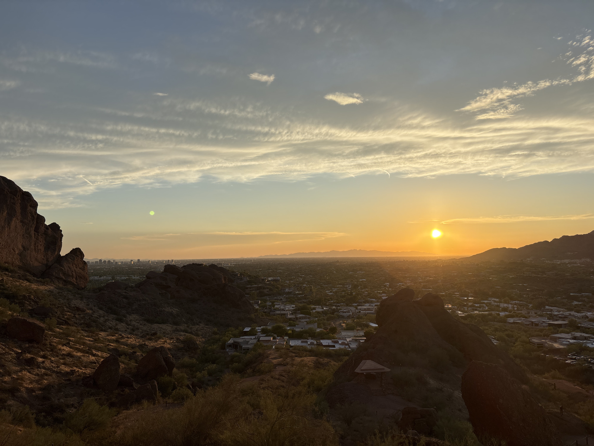 The image depicts a panoramic sunset view from a mountainous vantage point overlooking the city of Phoenix, Arizona, in the United States of America. The sun, a bright orange orb, is setting on the right side of the frame, casting a warm glow across the sky and landscape. Wispy clouds streak across the upper portion of the sky, tinged with orange and yellow from the setting sun.
In the foreground, rugged, rocky terrain with sparse desert vegetation dominates the left and bottom portions of the image. Large boulders and rock formations are visible, with shadows creating a sense of depth. A small, open-air pavilion with a pyramidal roof is situated on the right side of the foreground, with steps leading up to it.
Beyond the immediate rocky foreground, the landscape transitions into a sprawling urban environment. Numerous residential buildings, characterized by their flat roofs and a mix of architectural styles, are visible, interspersed with green trees and vegetation. The city extends into the distance, eventually merging with a hazy horizon where faint mountain ranges can be seen. The overall scene is characterized by the warm, golden light of sunset illuminating the natural desert landscape and the urban sprawl of Phoenix. There are no people clearly visible in the image.