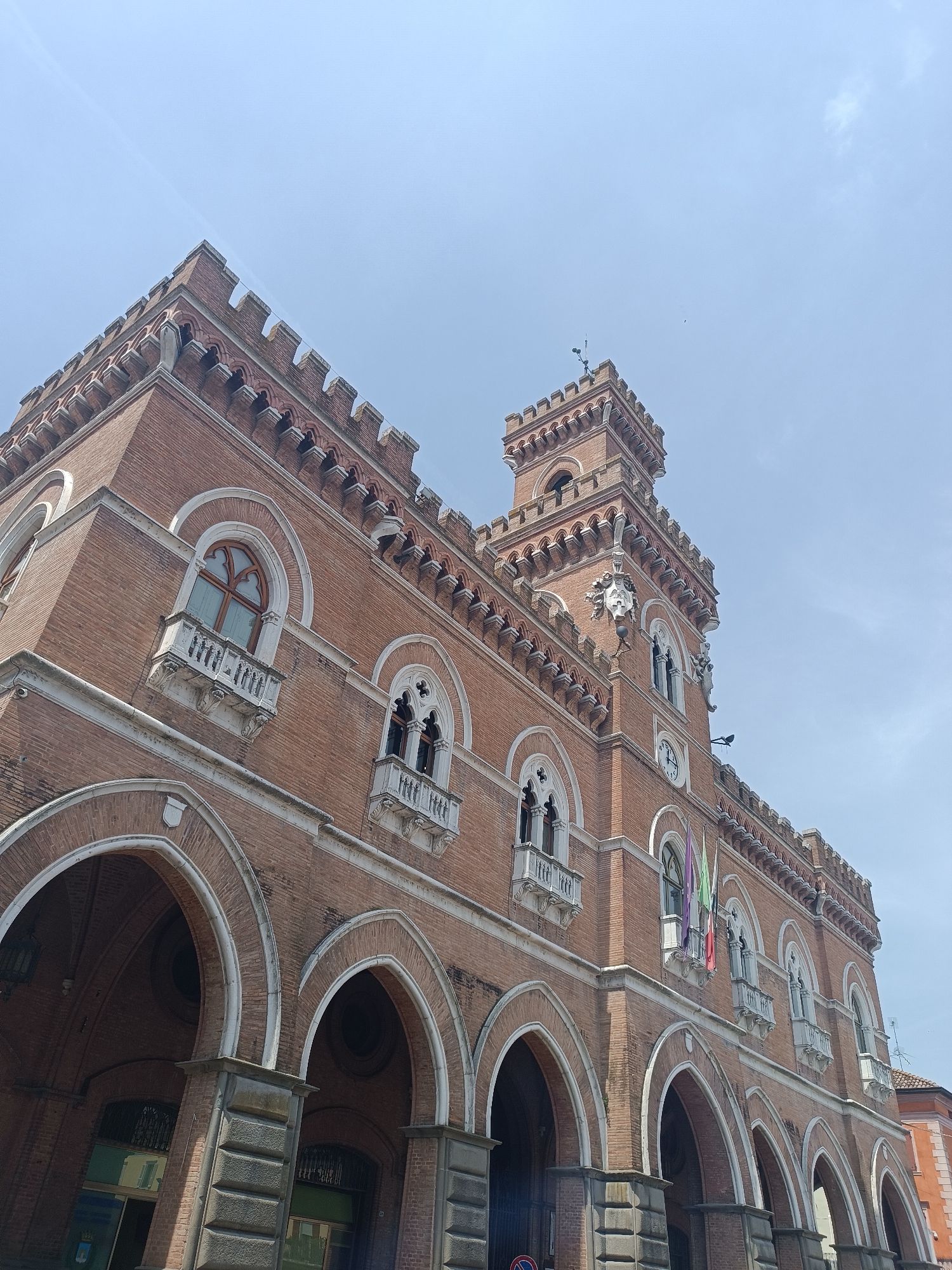 The image shows the exterior of a red brick building with arched entrances and windows, a tower with a clock, and flags. The architectural style appears to be Gothic or Neo-Gothic. The sky is clear and blue with a few wispy clouds. The building is likely a public building such as a town hall or a museum.
The building depicted is the Palazzo Comunale (Town Hall) of Casalmaggiore, Italy. The specific architectural features visible, including the crenellations along the roofline, the arched loggias at the ground level, the decorative windows with tracery, and the prominent tower with a clock, are characteristic of this building. The flags shown are likely the Italian flag and the flag of the Lombardy region, given the building's location. The bright, clear sky suggests the photograph was taken on a sunny day. There are no people visible in the image.