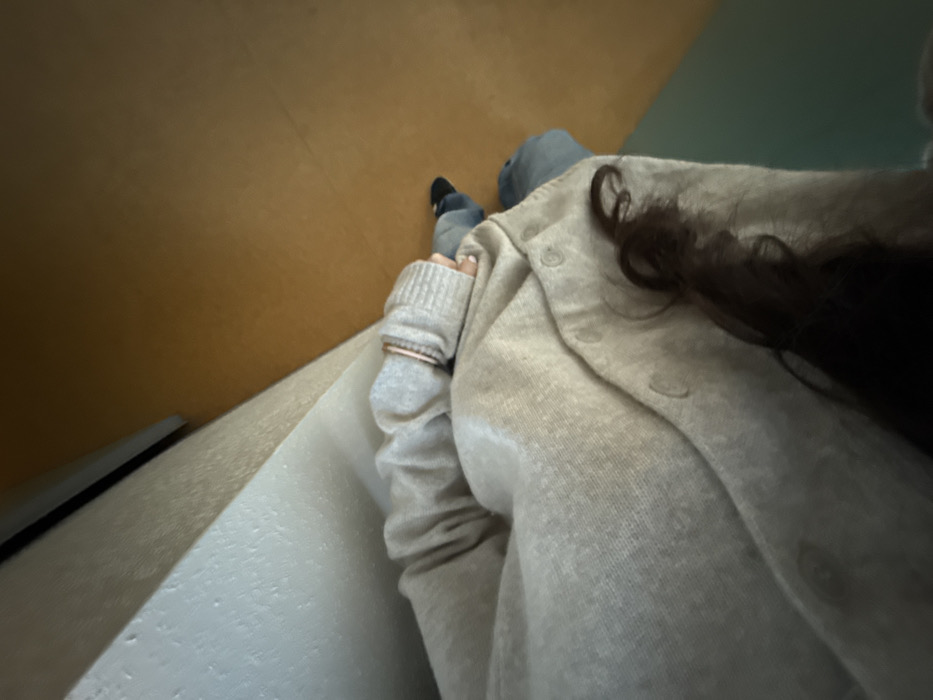 A person wearing a grey sweater and jacket sits on a white, cushioned surface. Their dark hair is visible on the right side of the frame. The background consists of a tan wall on the left and a green wall on the right. A black object is seen near the bottom left corner. The person is wearing a ring on their left hand, which is visible in the foreground. Part of their feet are visible in the center of the frame.