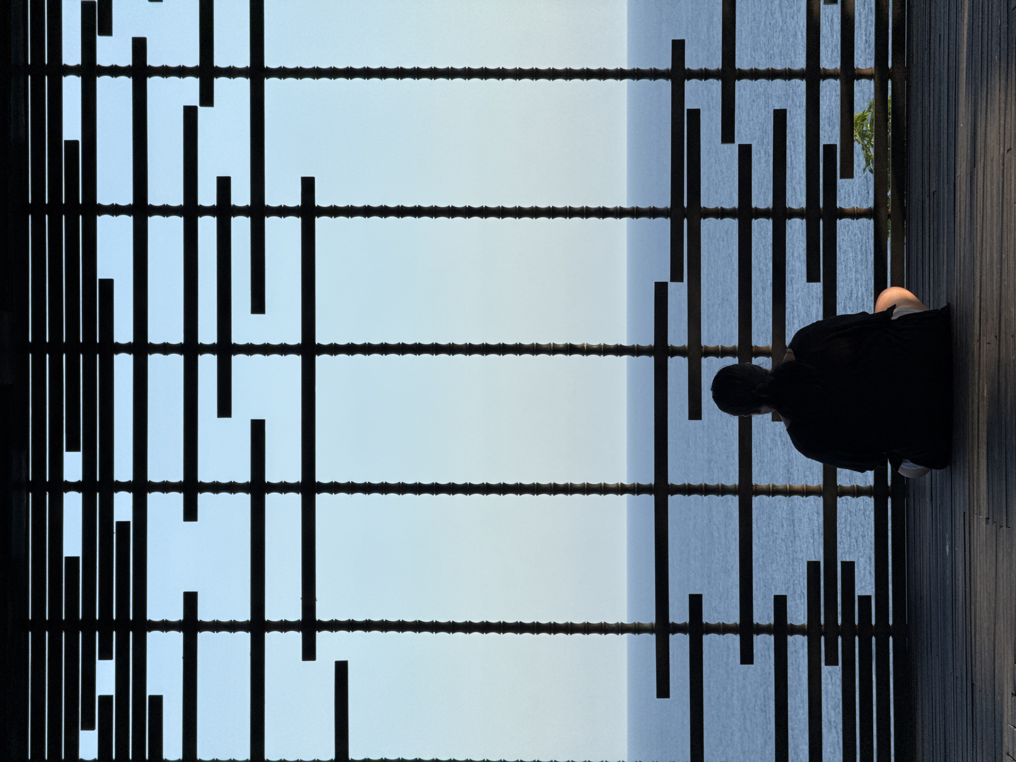 A person sits alone near a latticed fence on a wooden deck facing a body of water, possibly the ocean. The scene is dominated by the dark, vertically oriented latticework. The person is dressed in dark clothing, and their form is partially obscured by the lattice. The wooden deck runs parallel to the water, and a hint of green foliage is visible in the upper right corner. The location is Kuta Selatan, Indonesia.