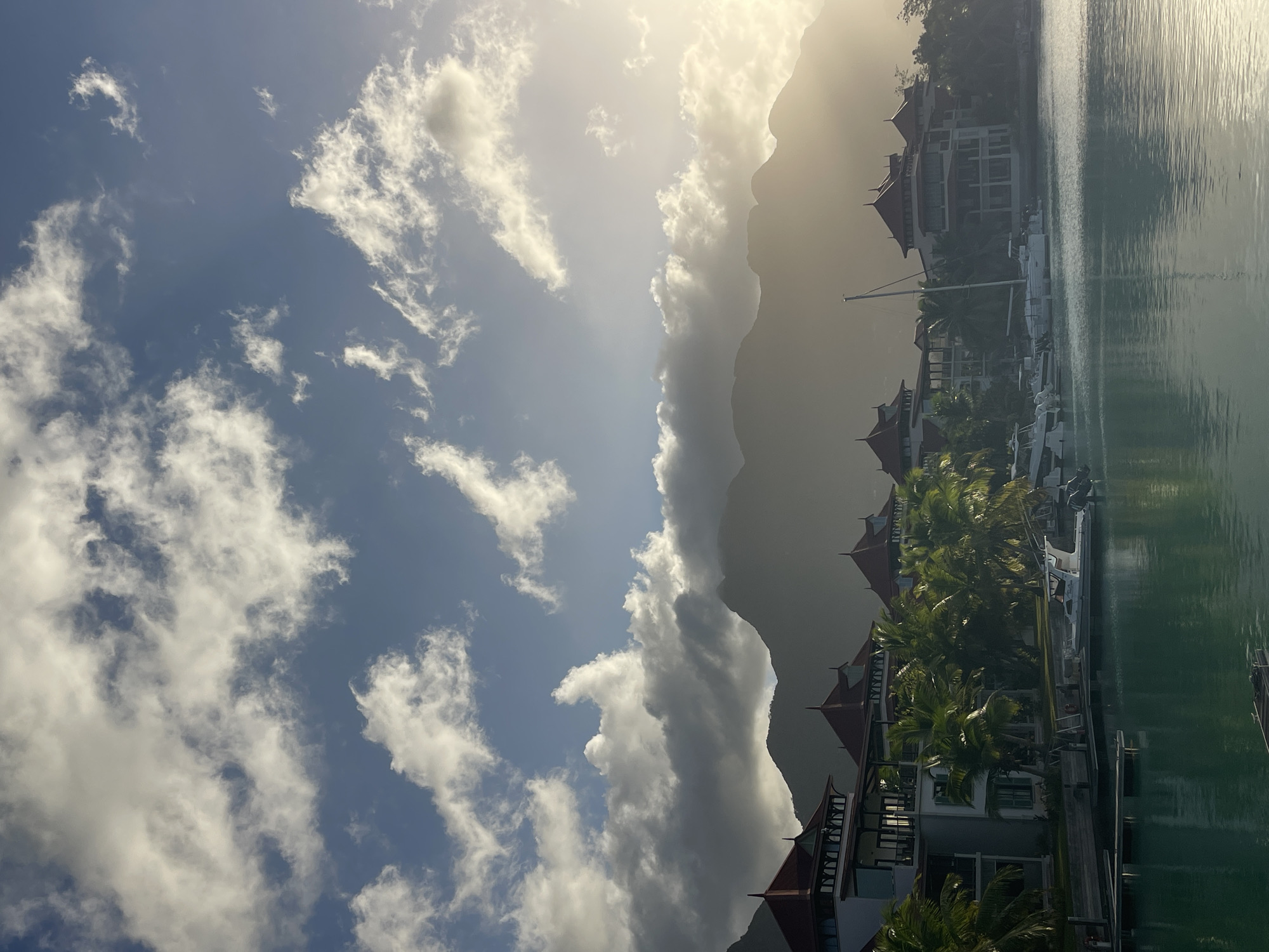 The image depicts a waterfront view of luxury resorts with distinctive red-tiled roofs, situated in the city of Providence, Seychelles. Palm trees line the waterfront, separating the buildings from the tranquil, turquoise water of the harbor. Several boats, including a sailboat and smaller motorboats, are docked. In the background, imposing, mist-shrouded mountains rise, silhouetted against a dramatic sky filled with a mix of bright sunlight breaking through scattered clouds and darker, heavier cloud formations. The overall scene conveys a sense of tropical serenity and upscale tourism. There are no people visible in the image.