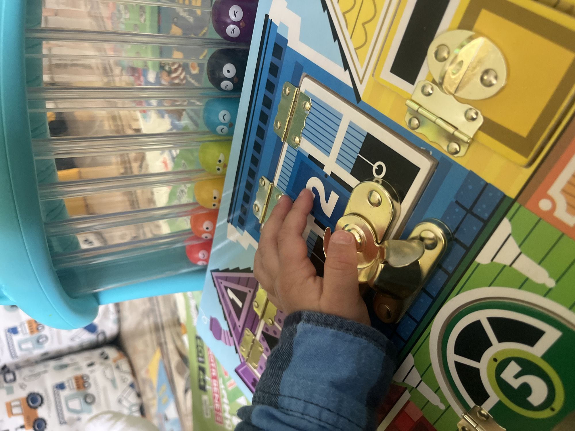 This indoor image, taken in Onokivtsi, Ukraine, captures a child's hands-on engagement with various toys during the daytime. The primary subject is a young child's hand, partially visible in a blue long-sleeved shirt, actively manipulating a golden latch on a vibrant "busy board."
The busy board is designed with a house or building theme, featuring multiple colored sections, golden hinges, and various latches and fasteners. Visible numbers on the board include "2" on a blue section, "1" on a purple section, and "5" on a green section, suggesting a learning or matching component. The child's hand is focused on unlocking or fastening one of the latches, indicating an activity involving fine motor skills and exploration.
Adjacent to the busy board, on the left side of the frame, is another toy. This consists of a bright blue plastic frame holding several clear, horizontal tubes. Inside these tubes are multiple small, round, colorful figures, each with a simple, cartoon-like face. The visible colors of these figures include purple, black, blue, yellow-green, yellow, and orange/red. This toy appears to be a "drop and roll" or sensory activity for the figures.
In the lower left background, a patterned fabric is visible, possibly part of a cushion or blanket, decorated with illustrations of construction vehicles like trucks and excavators. Other background elements are blurred, suggesting a typical home environment with other items or furniture. The lighting is bright and even, indicative of natural daylight indoors. There are no specific external location cues for Onokivtsi, Ukraine, beyond the context provided. The scene conveys a sense of focused, exploratory play.