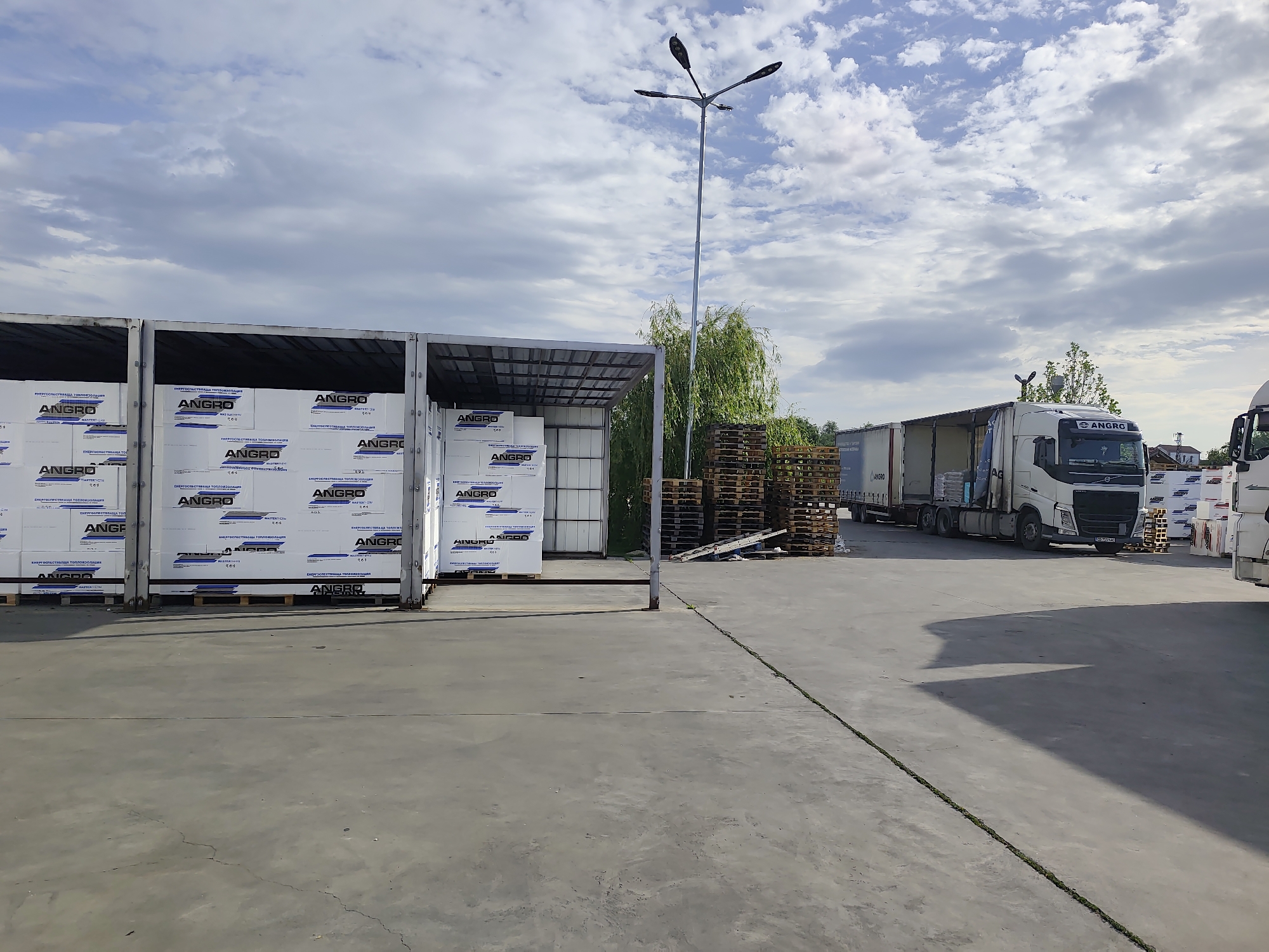 Here is a forensic description of the image:
**Location:** An outdoor industrial area in Grigorevo, Bulgaria, identified by the signage on the materials.
**Scene:** The scene displays a warehouse or storage yard filled with construction or insulation materials, identifiable by the "ANGRO" logo and descriptions (in Bulgarian) related to energy-saving thermal insulation on the packaging. These materials are stacked on pallets within open-sided storage units.
Several stacks of empty wooden pallets are visible in the background. A large white semi-truck is parked in the distance, and a second one, only partially visible, is closer to the foreground.
**Objects:**
* Palletized stacks of materials labeled "ANGRO"
* Open-sided storage units
* Wooden pallets
* Two semi-trucks
* A tall, modern street light
* Concrete pavement with expansion joints
**Environment:** Overcast sky with patches of blue, suggesting daylight conditions. The area appears to be a commercial or industrial zone.