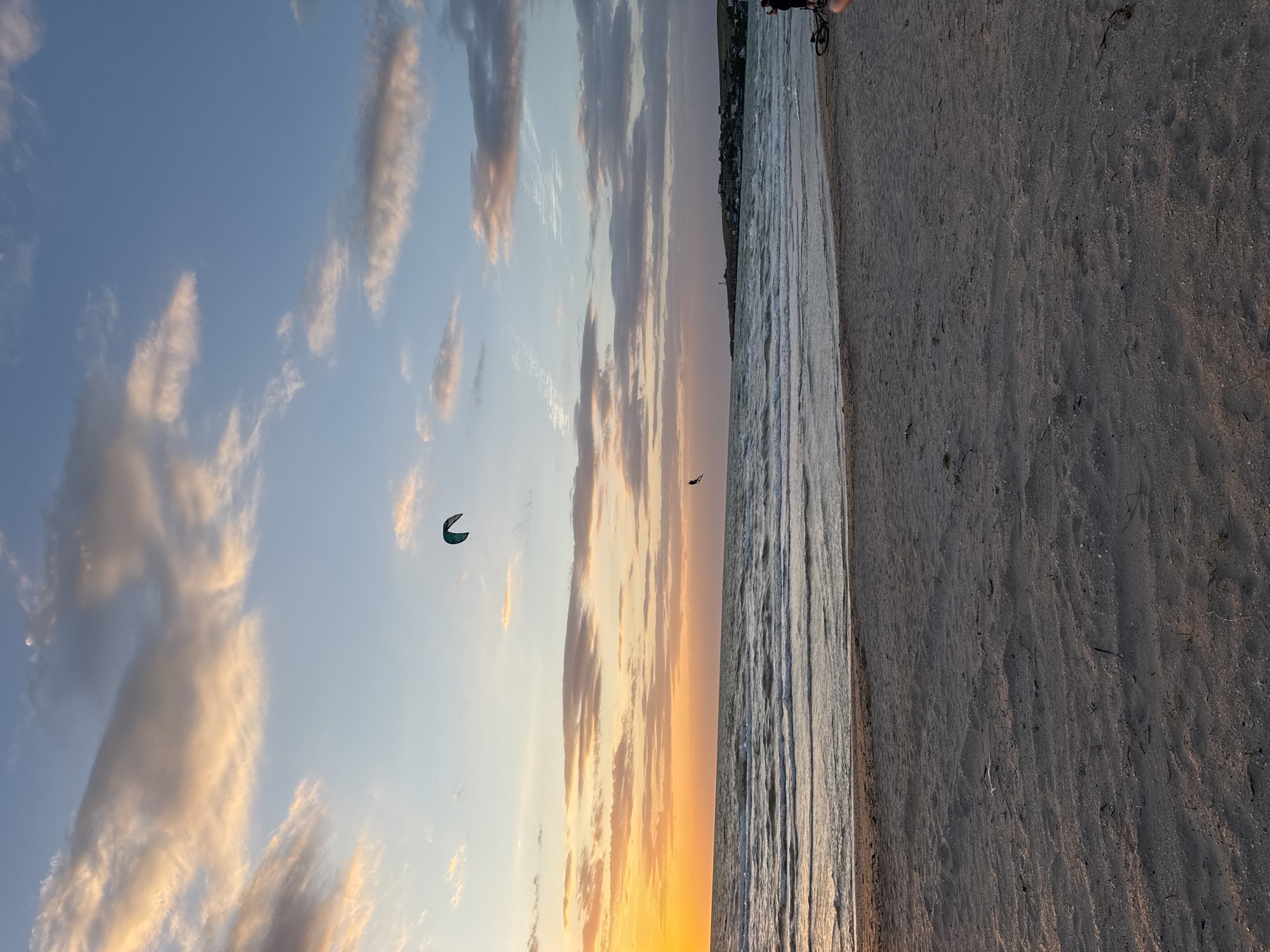 The image shows a beach at sunset. The beach is on the right of the frame, with the ocean to its left. In the distance, across the water, is a line of buildings. The sky is filled with clouds tinted in orange, yellow, and pink by the setting sun. There is a kite flying high in the sky. In the upper right corner of the frame, a person is seen riding a bicycle. The location of the city is Unknown, Unknown.