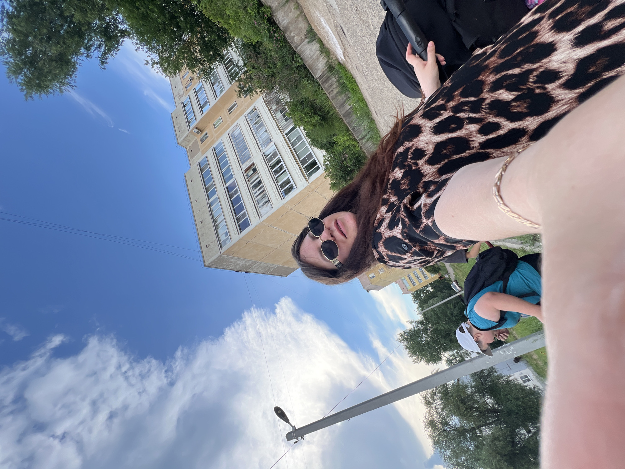 A wide-angle, selfie-style photo depicts a woman with long brown hair and sunglasses, wearing a leopard-print dress. She is holding a stroller handle with her right hand and has a gold bracelet on her left wrist. A boy wearing a blue shirt and a white cap with a black backpack walks behind her. In the background, a tall, light-brown apartment building with multiple balconies is visible, along with trees and a street lamp. The scene appears to be in an urban environment, likely a residential area in Noviy, Russia, based on the architecture and the presence of a stroller and child. The sky is bright blue with scattered white clouds.