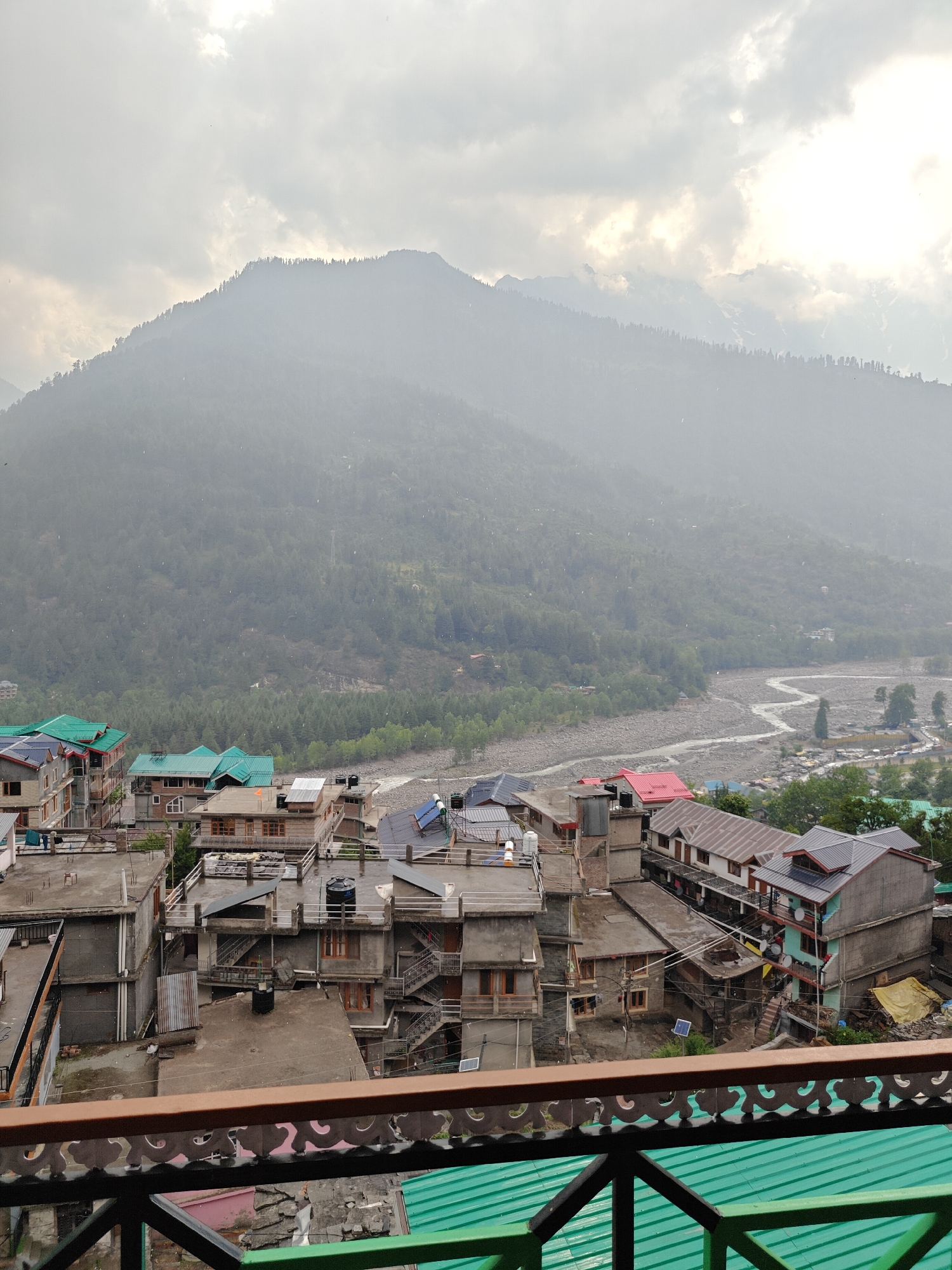 This image, taken from an elevated perspective, captures a hazy view of Vashisht, India, a town nestled in the Himalayas. The scene is dominated by a large, mist-shrouded mountain range in the background, its slopes covered in dense green trees. A winding river snakes through the valley below, with a small settlement of buildings on its banks.
In the foreground, a cluster of multi-story buildings with flat roofs and varying architectural styles occupies the mid-ground. Many of these buildings have balconies and external staircases. Rooftops are adorned with water tanks, solar panels, and some appear to be undergoing repairs or construction, indicated by tarpaulin sheets. The predominant colors of the buildings range from muted grays and browns to some with brightly colored green and red roofs. The weather appears overcast, with a light rain visible as streaks falling through the air. The overall impression is of a bustling mountain town during a damp, cloudy day.