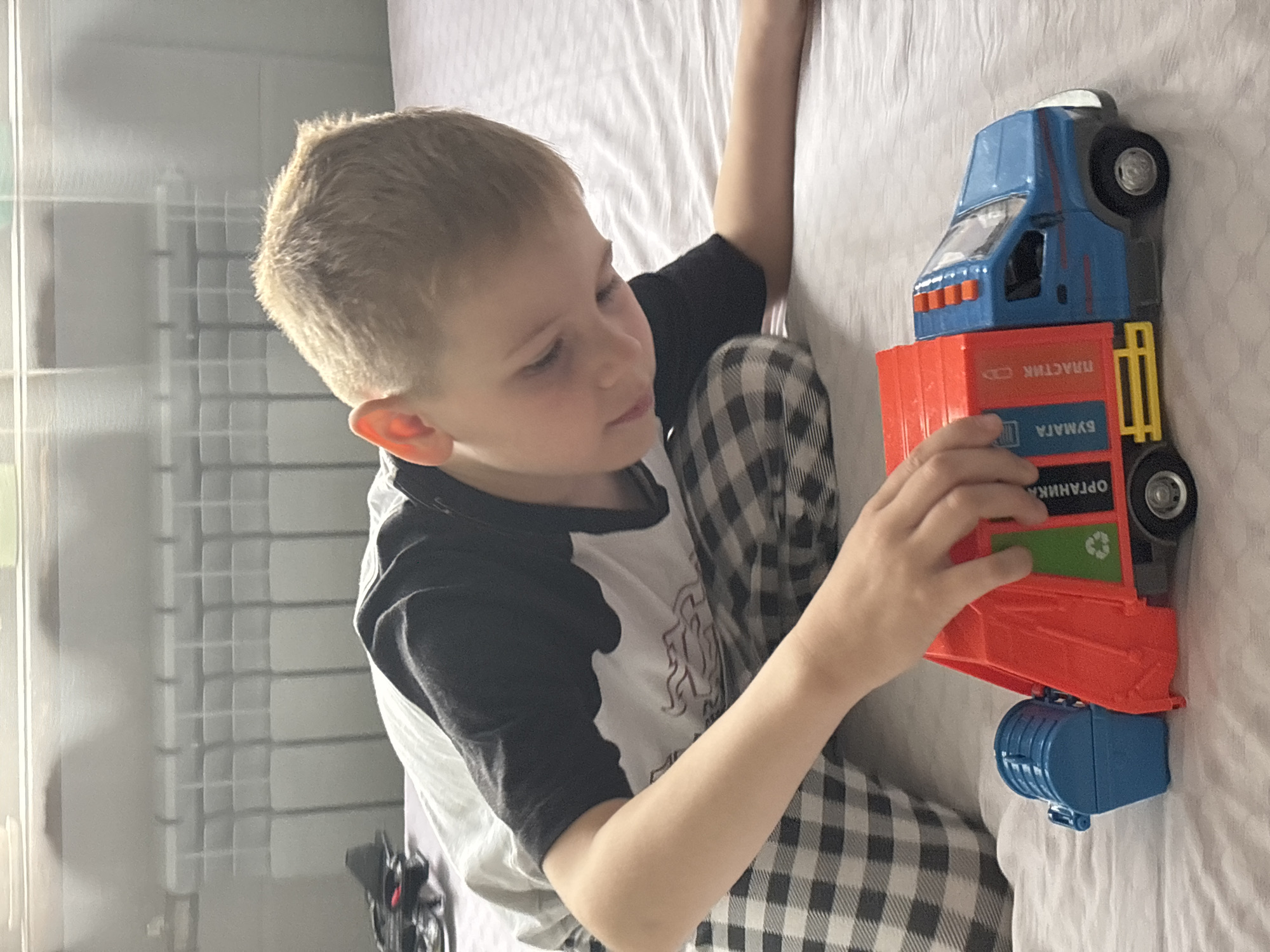 A boy is sitting on a bed and playing with a toy truck. The truck is blue and red and has labels in Russian. The boy is wearing a white and black shirt and checkered pants. The bed is covered in a white sheet. A window with a grid pattern is in the background. There is no definitive evidence to confirm the location as Fedorino, Russia.