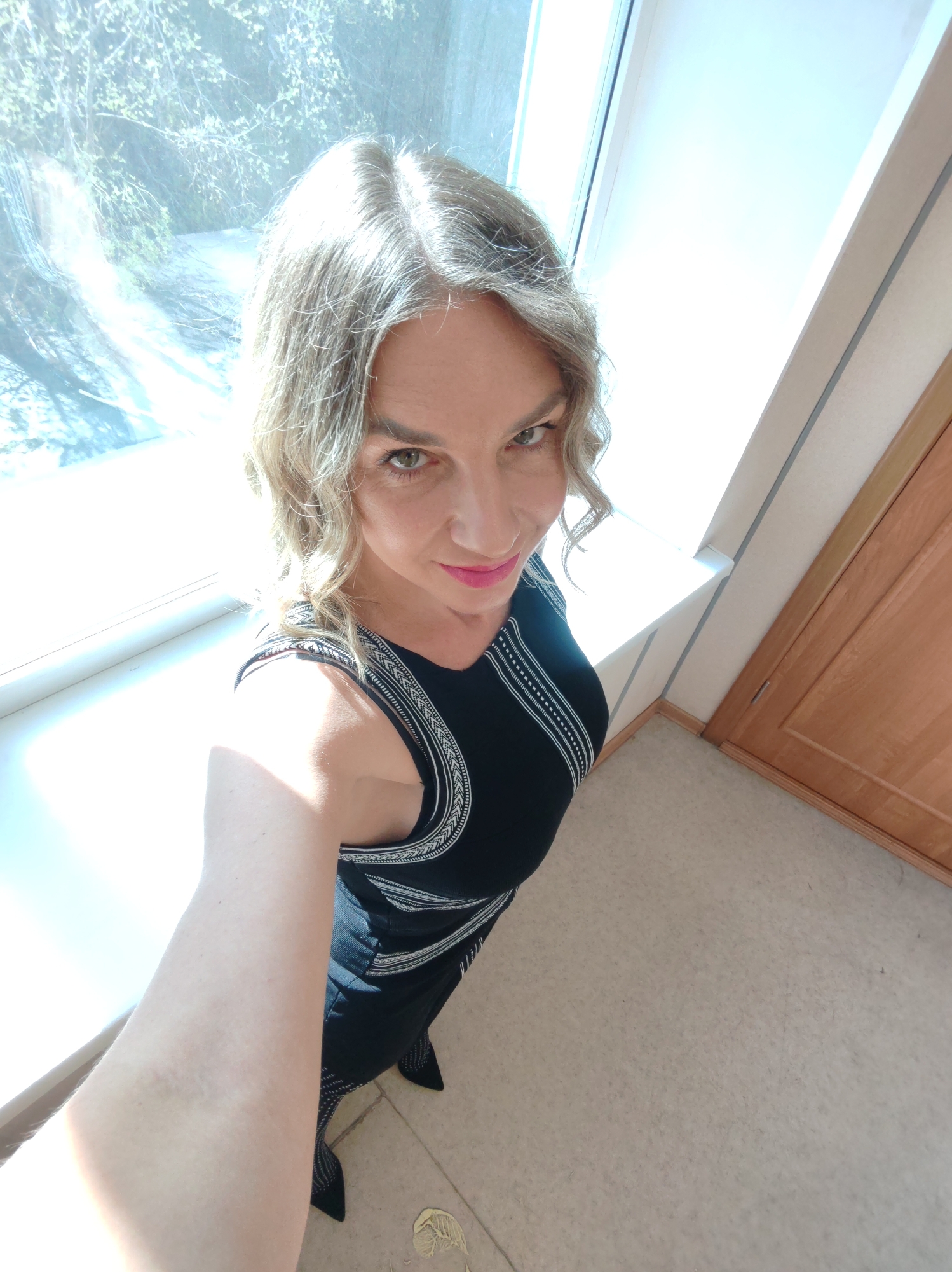 A high-angle selfie shows a woman with blonde curly hair, wearing a black dress with silver embellishments, standing inside a room. She is positioned between a window on her left and a closed door on her right. The window offers a view of trees and what seems to be a path. The room has a neutral-colored floor. The location in the city of Unknown, Unknown, is not ascertainable from the given image.