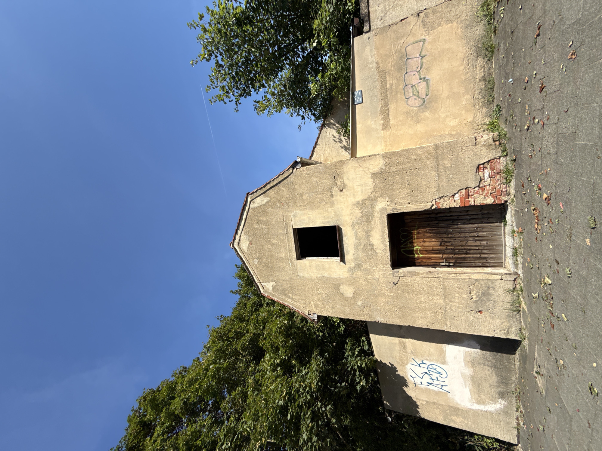 An eye-level, wide-angle shot captures the exterior of a weathered, tan building with graffiti on its walls, set against a bright blue sky with a faint contrail. The building, likely abandoned or in disrepair, features a peaked roof and a small, dark, empty window at the top. Below the window, a wooden door stands closed, adorned with some tagging. To the left of the door, a section of the tan wall has crumbled, exposing red brick. Graffiti in blue spray paint is visible on the lower left portion of the wall, and more graffiti in green is on the right side. Dense green trees flank the building on the left and right, partially obscuring the structure and the wall behind it. The foreground consists of a gray, paved surface littered with fallen leaves. The scene appears to be located in the city of Görlitz, Germany.