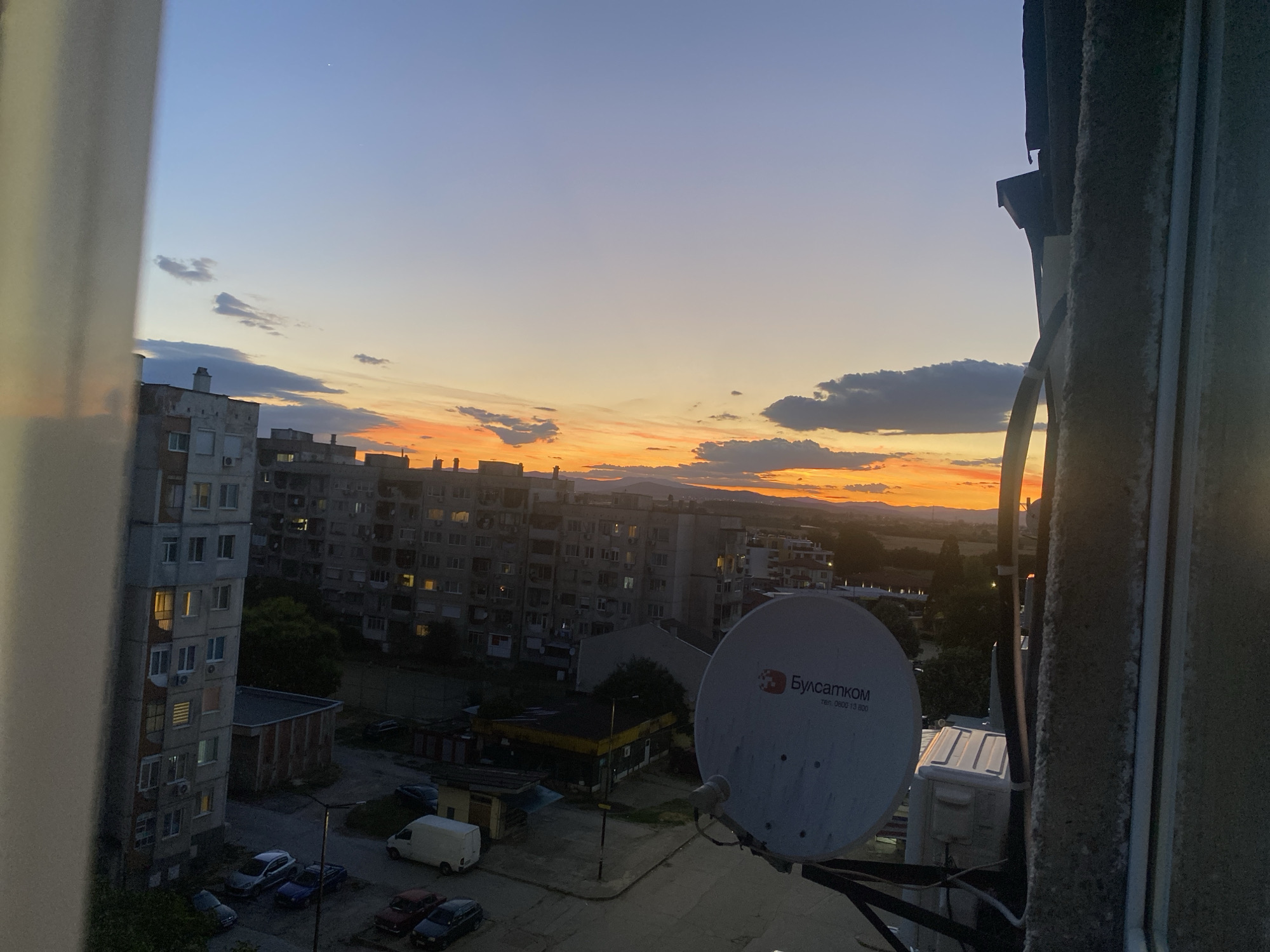The image is a telephoto view from a window, looking out at a city at dusk. The sky is a gradient of pale blue at the top, transitioning to orange and yellow at the horizon, with scattered clouds. In the foreground, a concrete pillar and window frame are visible on the right side, and a white curtain is on the left. Below the sky, a cluster of apartment buildings dominates the mid-ground. These buildings are multi-story, with many windows, some of which are illuminated. A satellite dish, with Bulgarian text that reads "Булсатком" (Bulsatcom) and a phone number, is prominently displayed on the right side of the image, positioned in front of the apartment buildings. Below the buildings, a street is visible with several parked cars, including a white van, a few sedans, and a pickup truck. A street lamp is also present. The overall setting appears to be a residential area within the city of Pazardzhik, Bulgaria. The lighting suggests the sun has recently set, casting a warm glow on the horizon. There are no people clearly visible in the image.