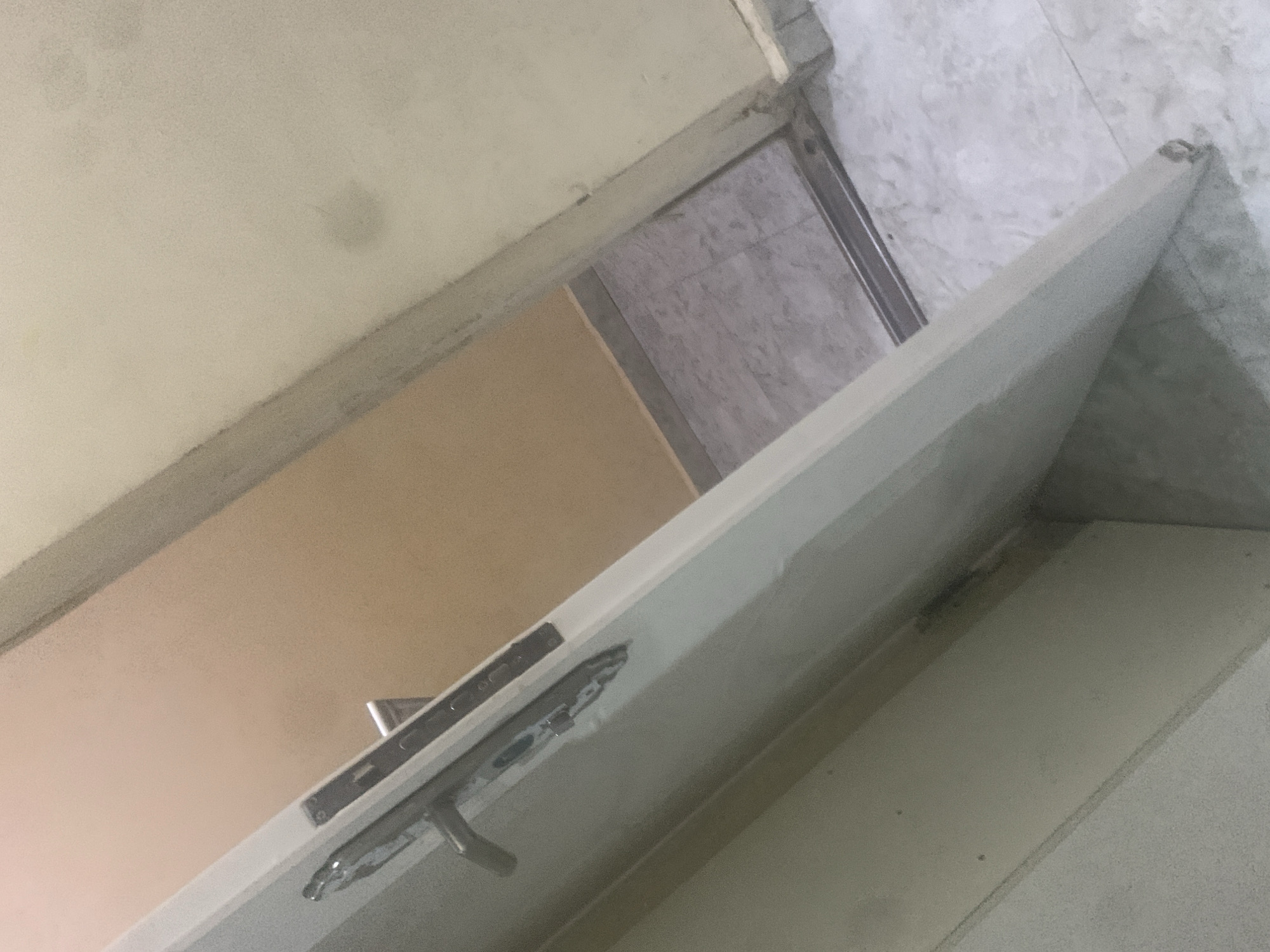 A slightly tilted, wide-angle interior shot shows a partially open white door with a silver handle and lock mechanism. The door is angled, revealing a peach-colored wall in the background and a floor with light gray tiles and what appears to be a silver-colored floor track. To the right of the door, a portion of a cream-colored wall with some faint smudges is visible. The scene is generic and could be any interior space with a door, therefore it is not possible to pinpoint a specific location in Jalingo, Nigeria.