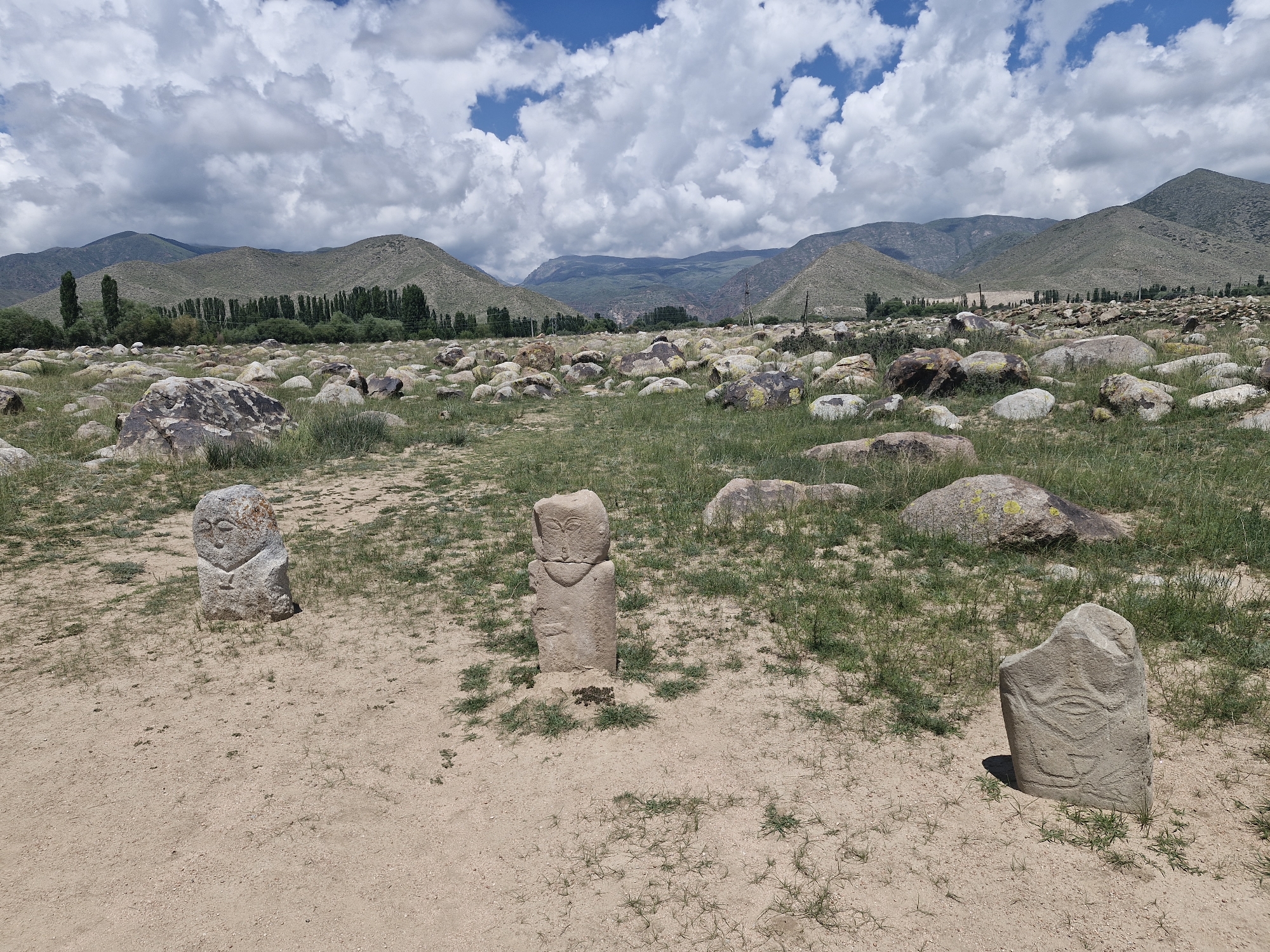 The image depicts a desolate, open landscape with scattered rocks and sparse vegetation under a cloudy sky. In the foreground, three stone stelae are visible. The stele on the left is roughly carved with what appears to be a face. The central stele is taller and more rectangular, also featuring a carved face with prominent eyes. The stele on the right is smaller and also displays carved features, resembling an eye and other abstract shapes. The background shows rolling, treeless hills and mountains. This scene is consistent with the area surrounding the Cholpon-Ata petroglyphs site in Cholpon-Ata, Kyrgyzstan, an archaeological site known for its open-air collection of ancient rock carvings. There are no people or animals visible in the image, and the environment appears dry and arid.