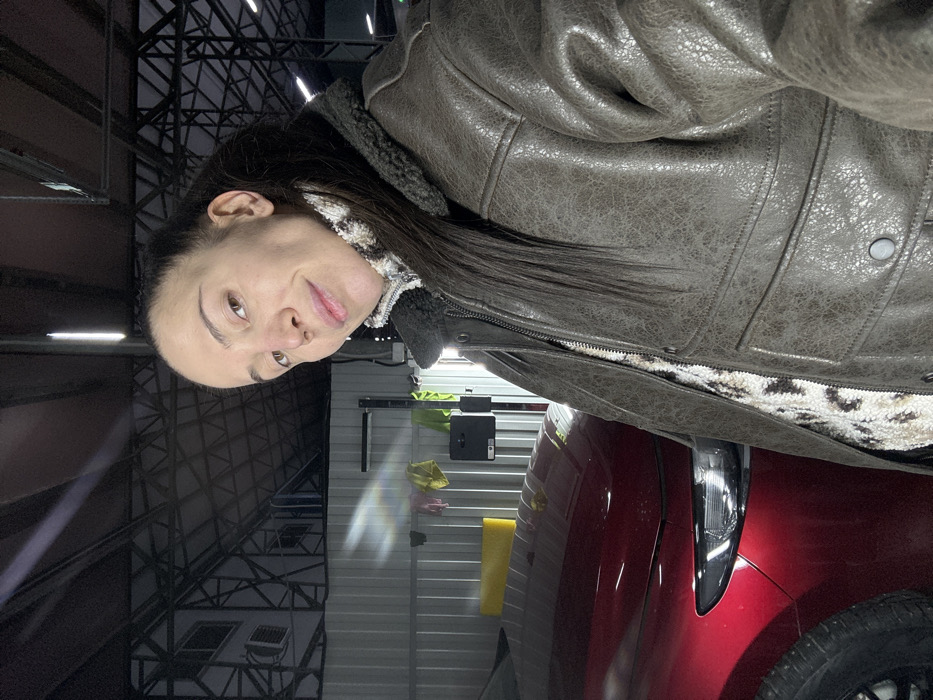A woman in a leather jacket stands beside a red car. She's wearing a patterned top with a fur-lined collar under the jacket. Behind her is a metal building with visible beams and artificial lighting. Cleaning supplies are visible against the wall. The woman has dark hair and is looking at the camera with a slight smile.