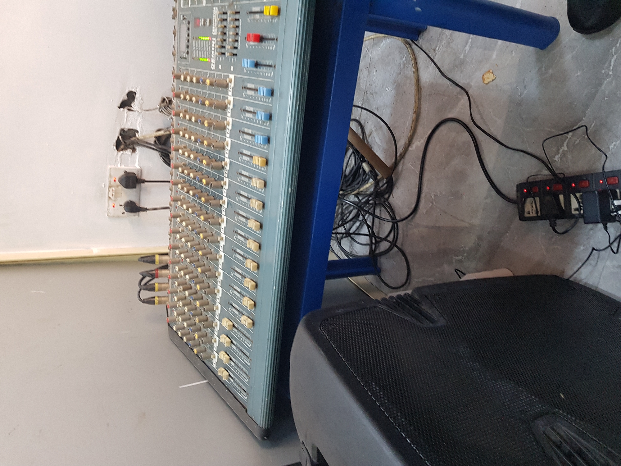 The image presents a scene in what appears to be an indoor space in Ikeja, Nigeria, based on the plug types visible. The scene consists of a grey wall with electrical outlets and a multi-plug adapter. Connected to the outlets and adapter are various cables and power plugs, suggesting a complex electrical setup.
A mixing console is positioned on a blue metal table, its surface covered with numerous controls, knobs, and sliders. Wires and connectors are visible, plugged into the side of the console. A black speaker is positioned near the console, partially visible in the lower portion of the image. The floor is a light grey color, marked by cable clutter and a small debris.