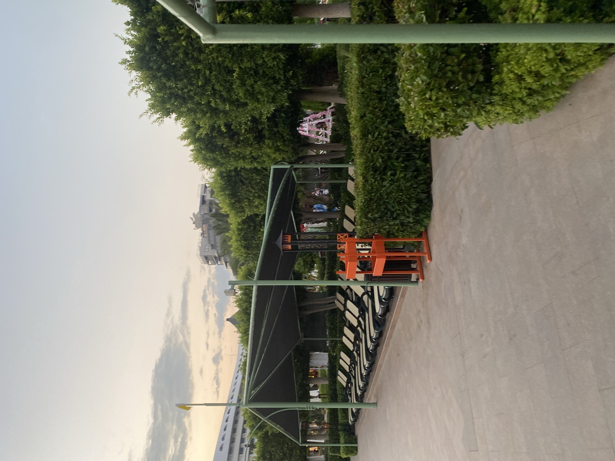 The image shows a resort area with a row of empty lounge chairs under a black canopy. In the foreground, a paved walkway is bordered by manicured green hedges. An orange metal structure, possibly a lifeguard stand, is positioned next to the lounge chairs. Behind the chairs and hedges, there are several large trees and buildings in the distance. The sky is overcast with soft, diffused light, suggesting it might be early morning or late afternoon. The overall scene depicts a tranquil outdoor recreational space in a warm climate, likely Aksu, Turkey, given the resort-like appearance and vegetation.