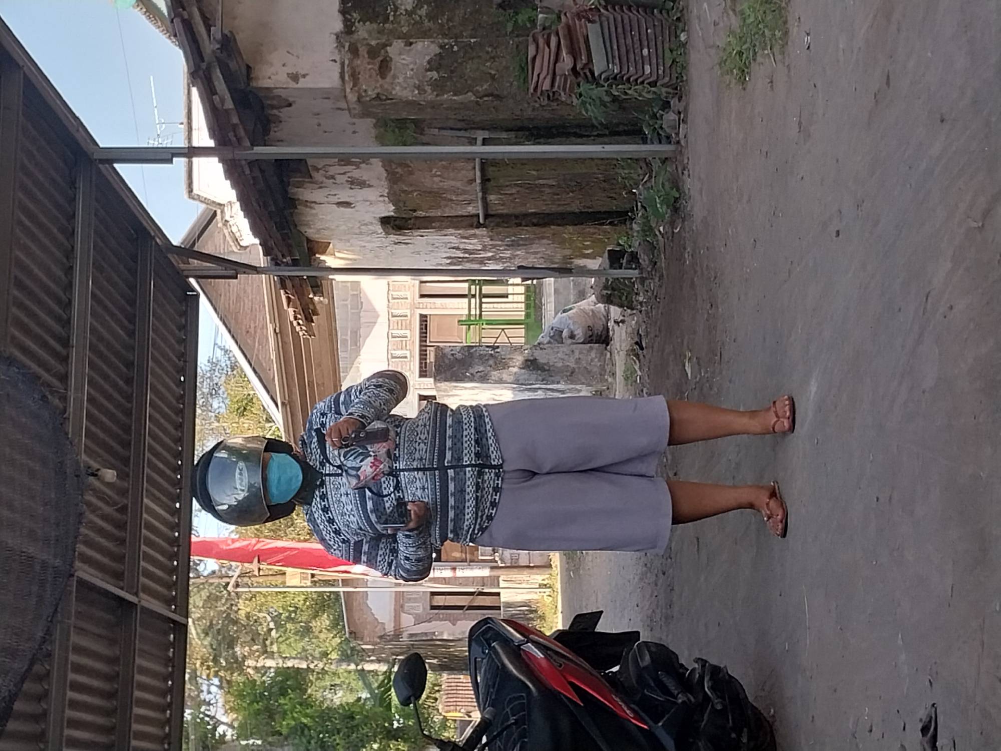 A person wearing a blue face mask and a helmet stands outdoors under a metal-framed, corrugated metal roof. The individual is dressed in a patterned zip-up jacket and light gray shorts, with brown flip-flops on their feet. They hold what appears to be a cell phone in their left hand and a small bag or pouch in their right. To the left of the person is the front portion of a red and black motorcycle. The background shows a rural or semi-urban setting with a rustic building, some vegetation, and what looks like a stack of roof tiles. The ground is paved with asphalt or concrete. The scene is illuminated by natural daylight.